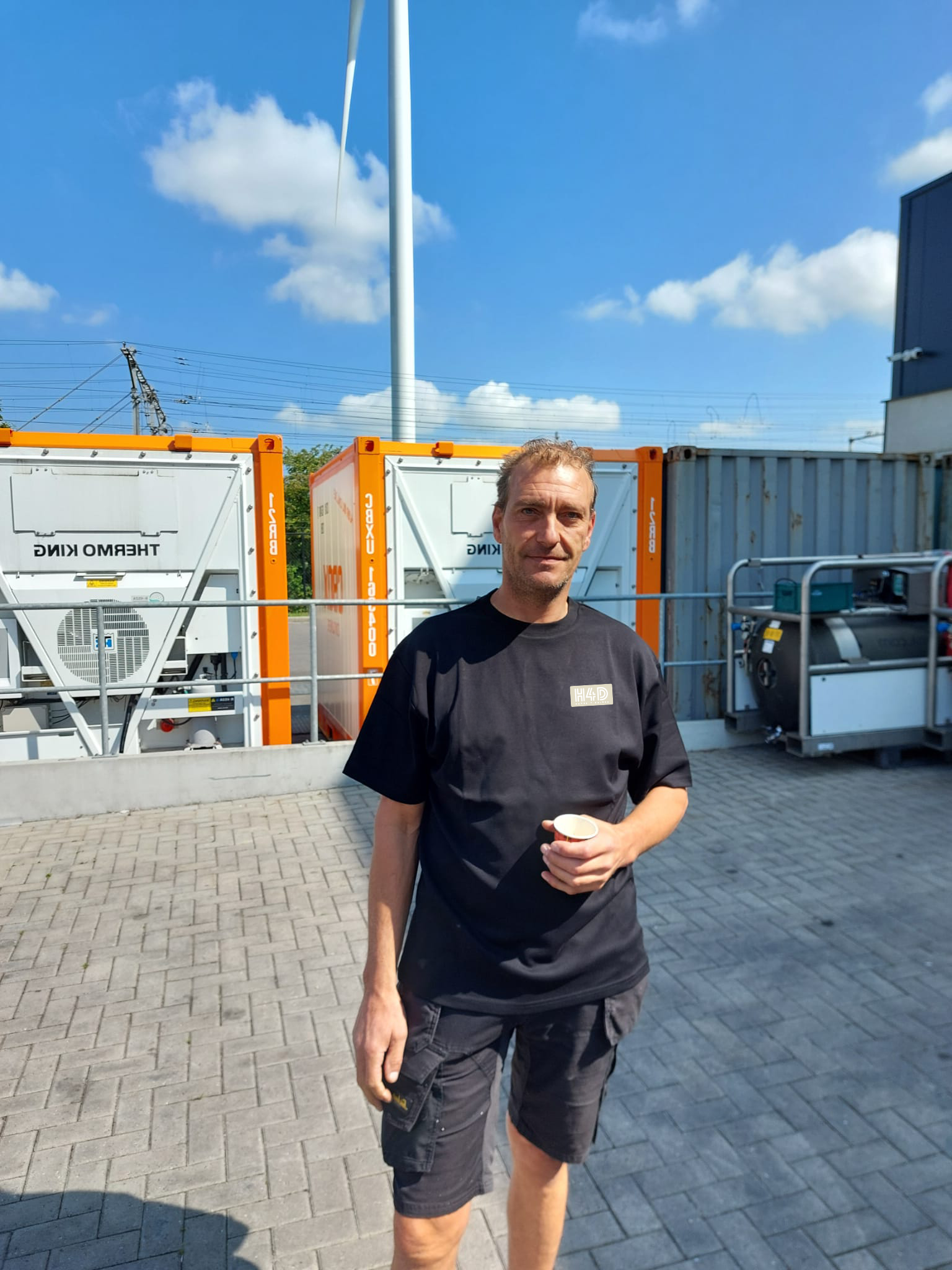 A man holding a small cup outdoors on a sunny day, standing on a paved surface with large equipment and a wind turbine in the background.