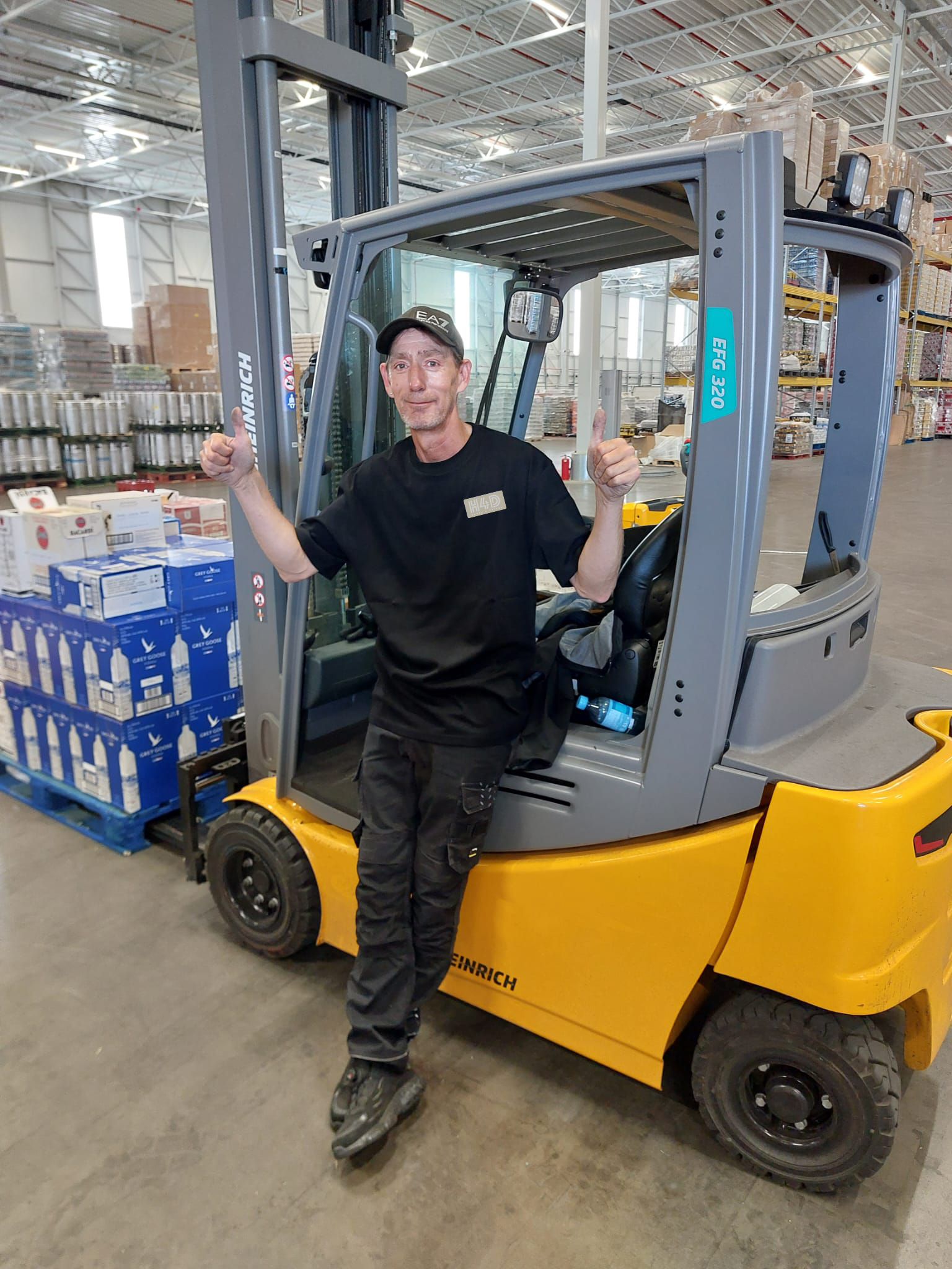 A man standing on the front fork of a yellow forklift inside a warehouse, giving a thumbs-up with both hands.