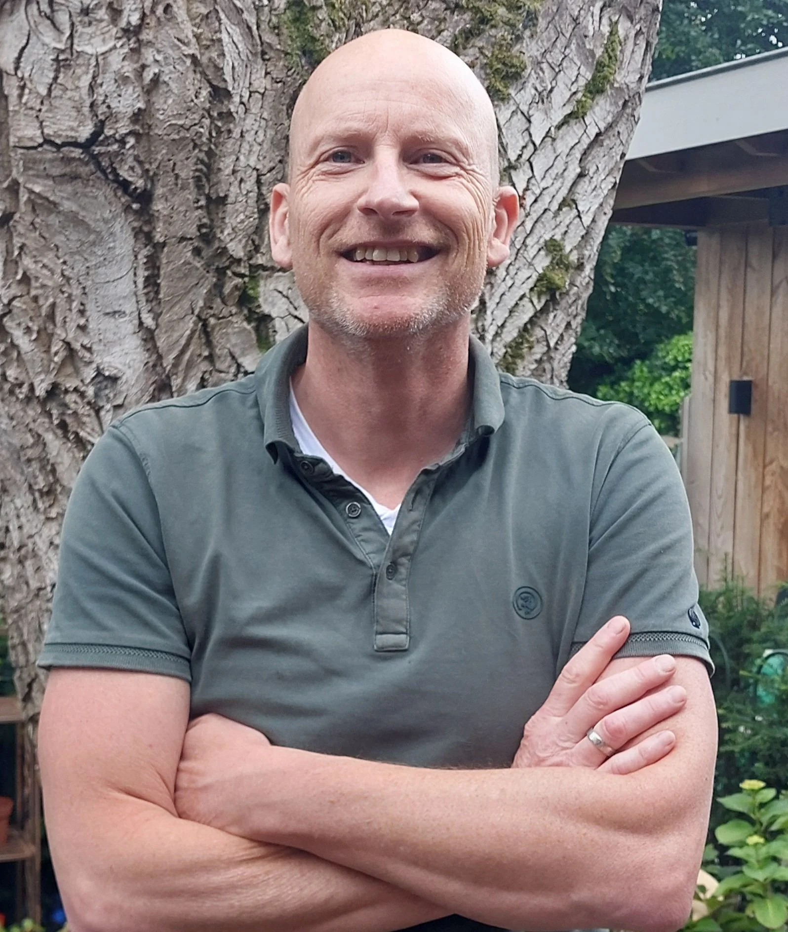 A smiling man with a bald head and light beard, wearing a short-sleeved, collared, dark green shirt, standing outdoors against a large tree trunk with green foliage and a wooden shed in the background.