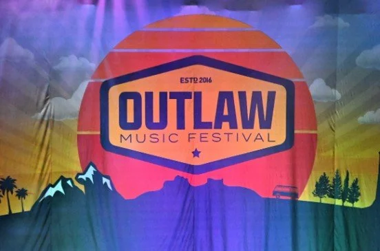 A colorful banner for the Outlaw Music Festival with mountains, trees, and a sunset background.