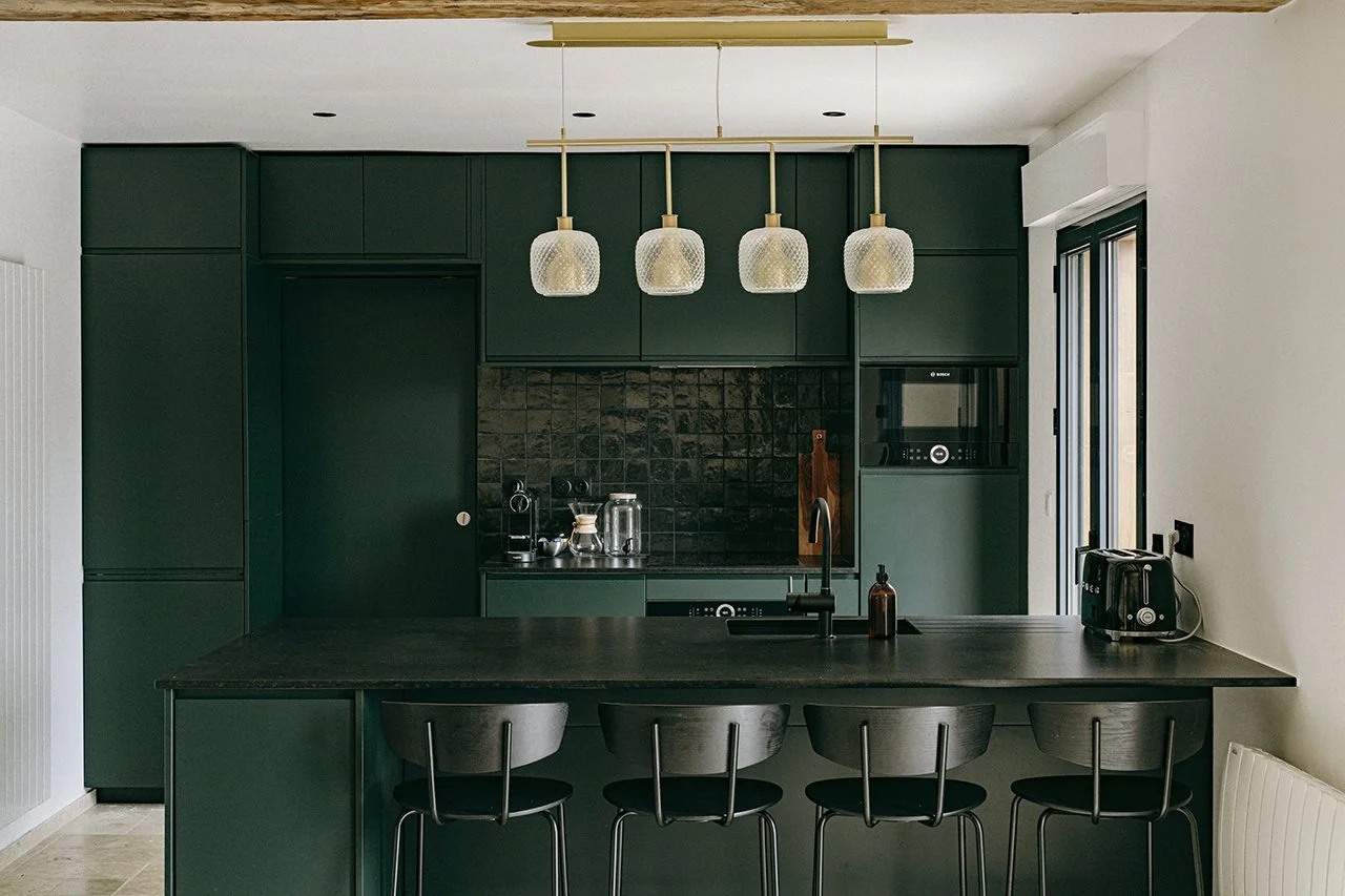 Cuisine moderne avec plan de travail noir, derrière des armoires vert foncé, éclairée par cinq lampes suspendues, avec un évier et un robinet, quelques appareils électroménagers dont un grille-pain et une cafetière, et un mur en brique noire.