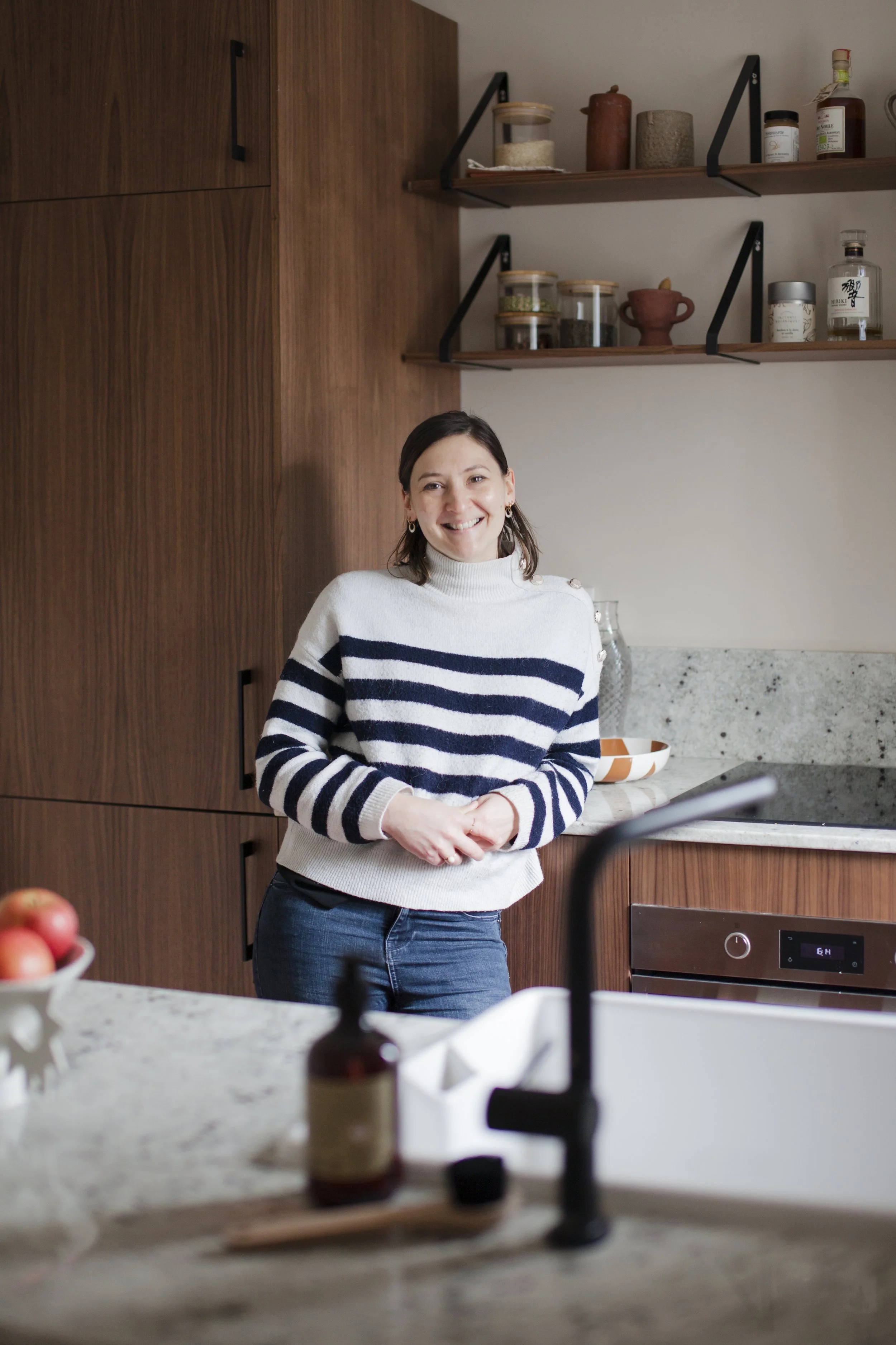 Une femme souriante avec un pull à rayures noires et blanches dans une cuisine moderne en bois et pierre.
