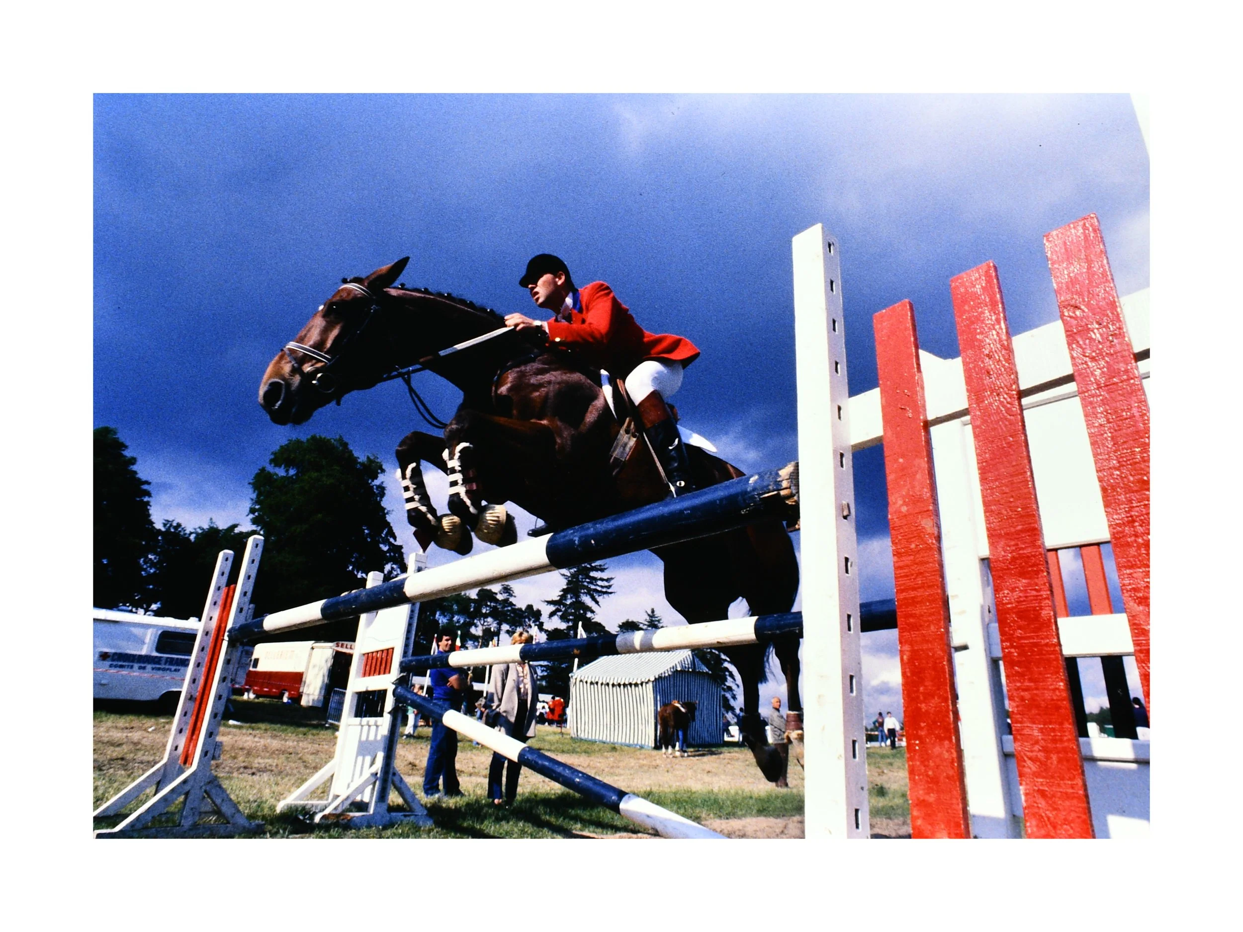 Photographie argentique d’un saut d’obstacle lors d’un concours hippique en 1985, cavalier en veste rouge franchissant une barre, archive de Didier Arrès.