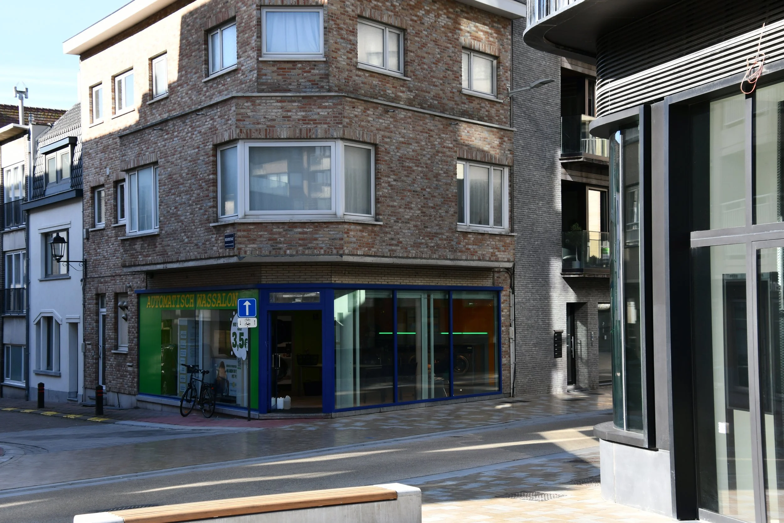 Een straat met een hoekgebouw dat een autowasbedrijf op de begane grond heeft, met een fiets vóór de ingang. De straat ligt nat, mogelijk door regen.