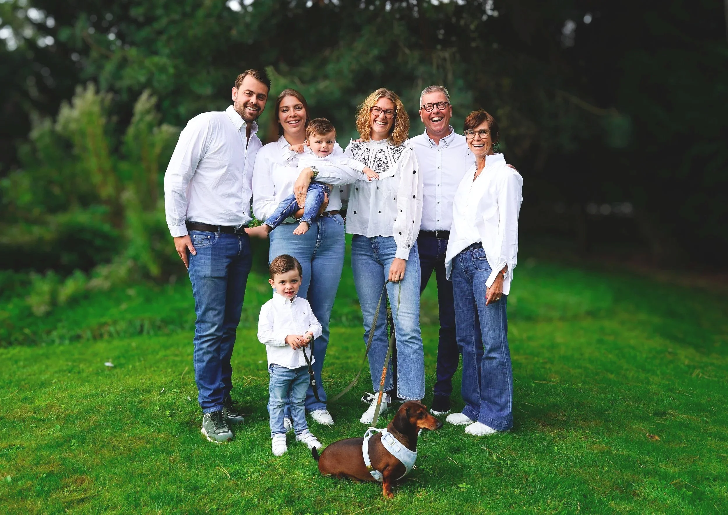 Gezin met vier volwassenen, twee kinderen en een hond poseren op een grasveld in de natuur, allemaal gekleed in witte shirts en jeans, lachend.
