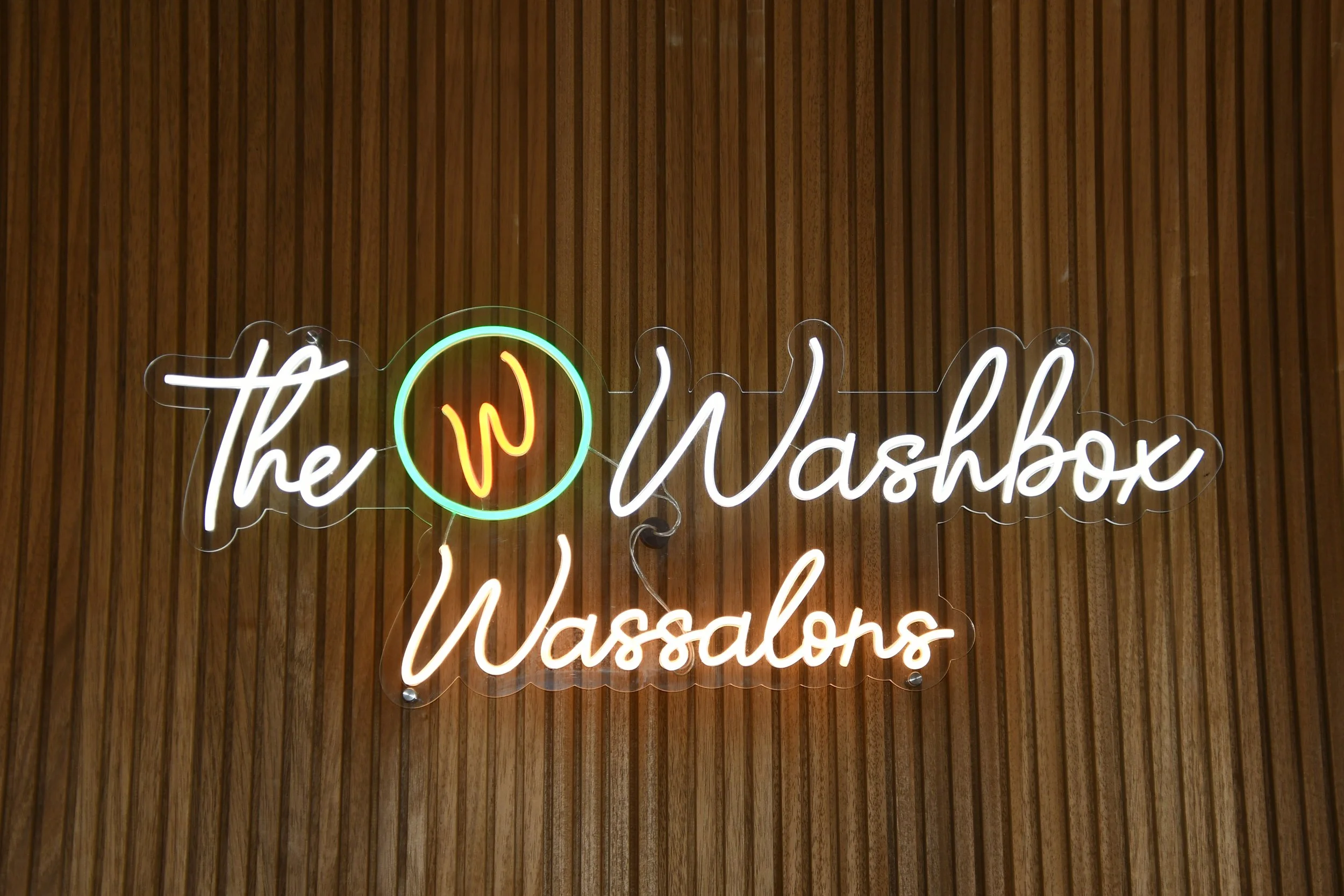 Neonbord met de tekst 'The Washbox Wassalon' tegen een houten wand.