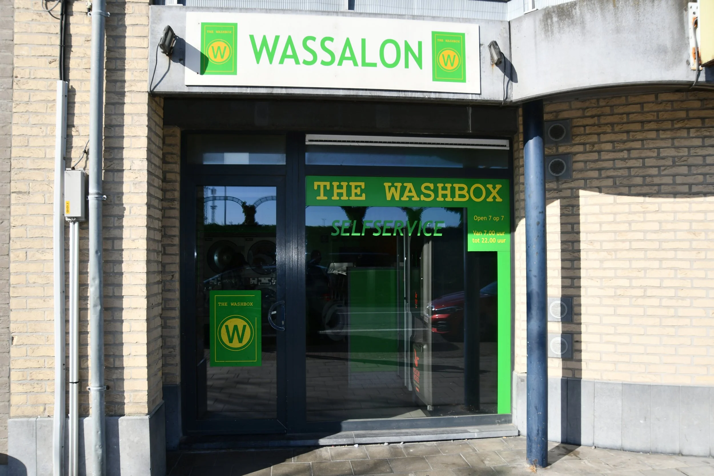 De gevel van een winkel met een witte en brick muur. Boven de ingang hangt een witte bord met groene tekst 'WASSALON' en twee groene logo's. Op de deur staat 'THE WASHBOX', 'SELF SERVICE', en openingstijden in het Nederlands.