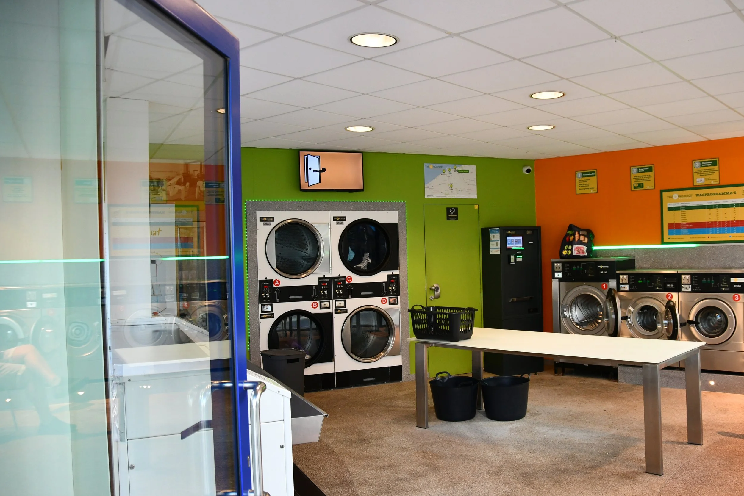 Binnenkant van een wasserette met wasmachines, droogautomaten, een tafel, en wasmiddelen.