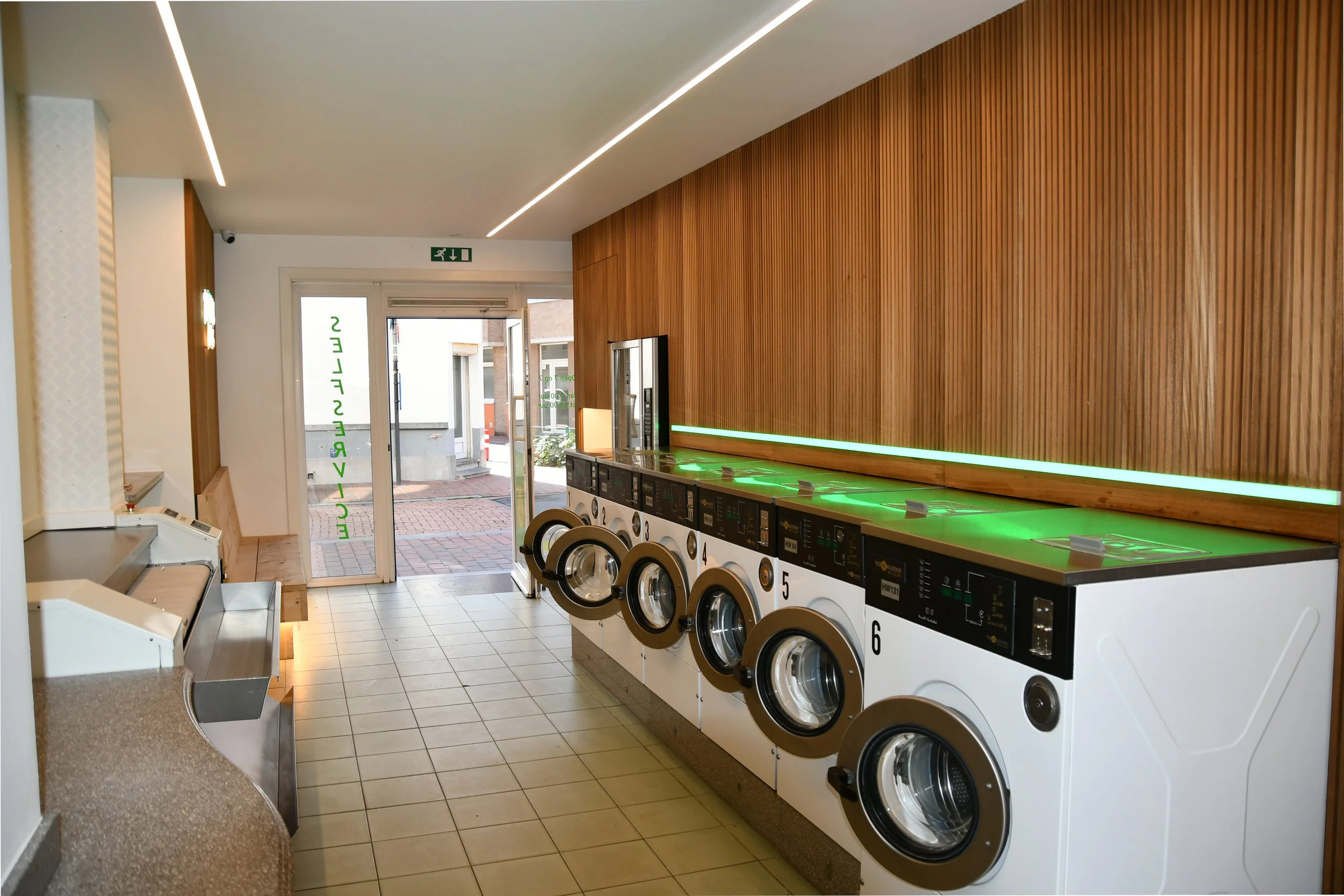 Wasruimte met wasmachines en wasdrogers, houten wand, betegelde vloer, deur met glas.