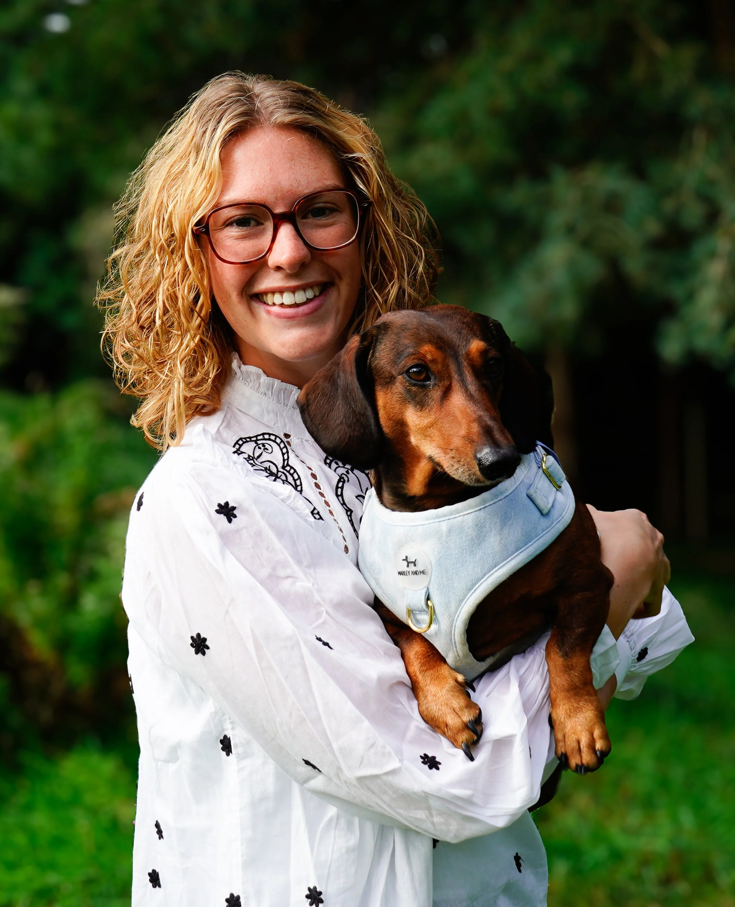 Lachende vrouw met bril houdt een dotje in haar armen buiten in een groene omgeving.