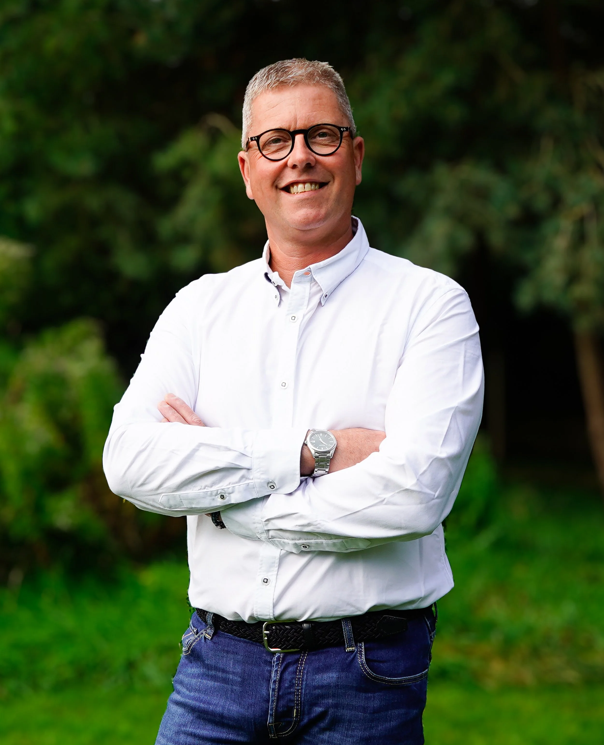 Man met kort, grijs haar en bril, lachend, gekleed in een witte blouse en blauwe jeans, staand met gekruiste armen in een tuin met groene bomen en gras.
