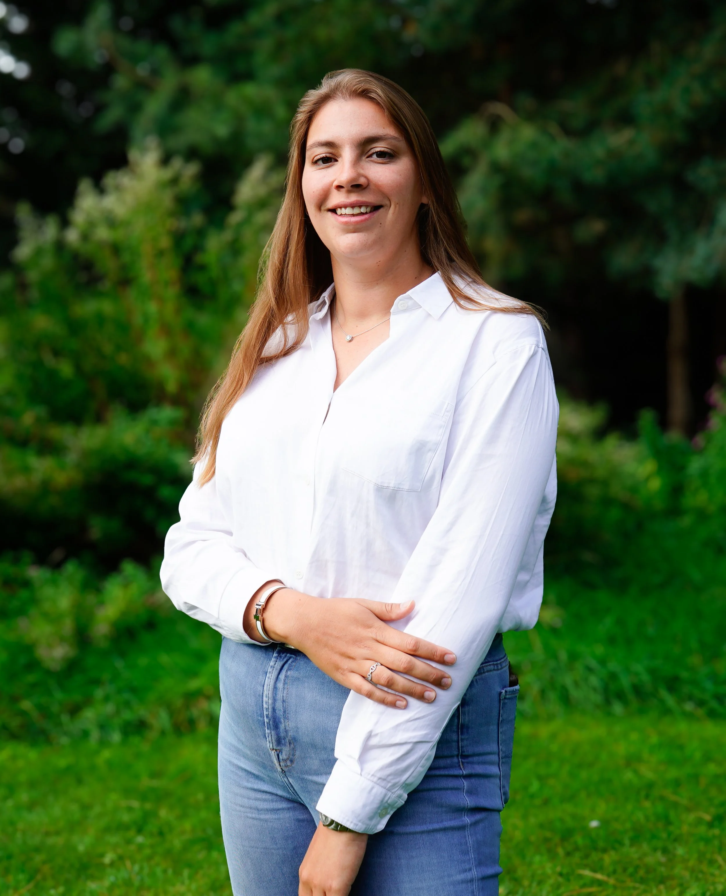 Portret van een jonge vrouw met bruin haar die in een tuin staat, lachend en gekleed in een witte blouse en blauwe jeans.