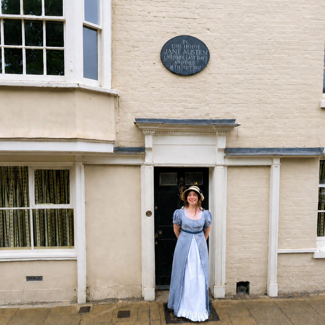 A woman dressed in historical clothing standing outside a beige brick house with a black door, a white doorframe, and windows with green curtains. There is a black plaque above the door stating it was Jane Austen's residence when she lived her last days, dying on July 18, 1817.