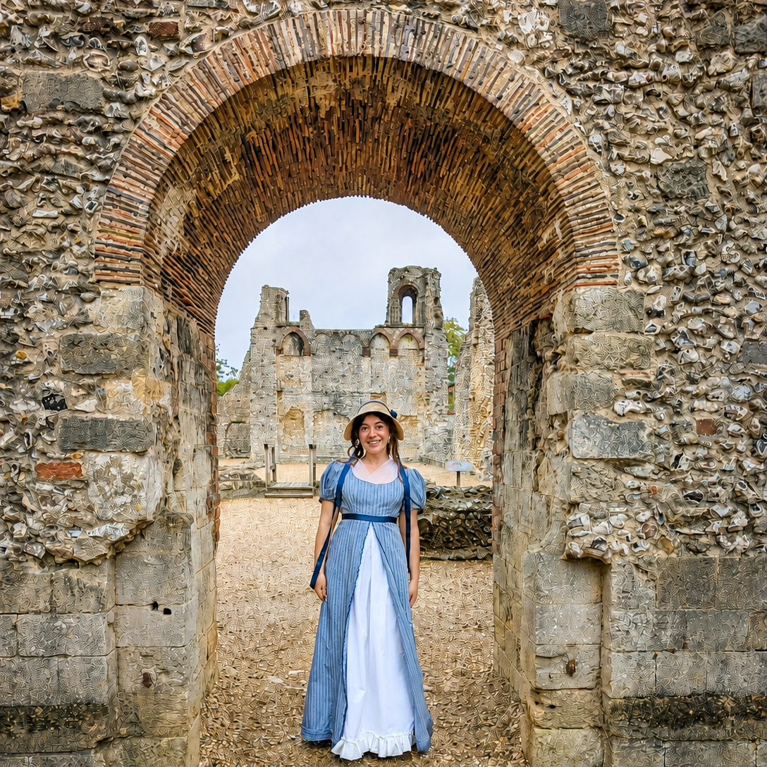 A woman in a blue and white dress and hat standing in an ancient stone archway with ruins in the background.
