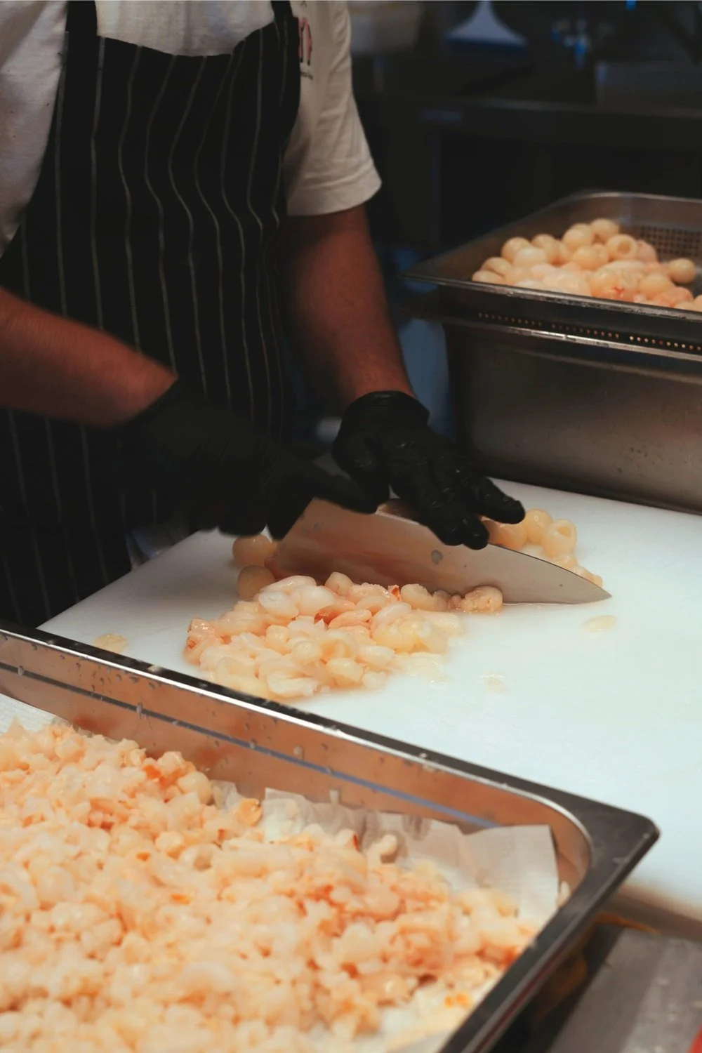 A chef wearing black gloves and a striped apron slices cooked shrimp on a white cutting board in a professional kitchen.