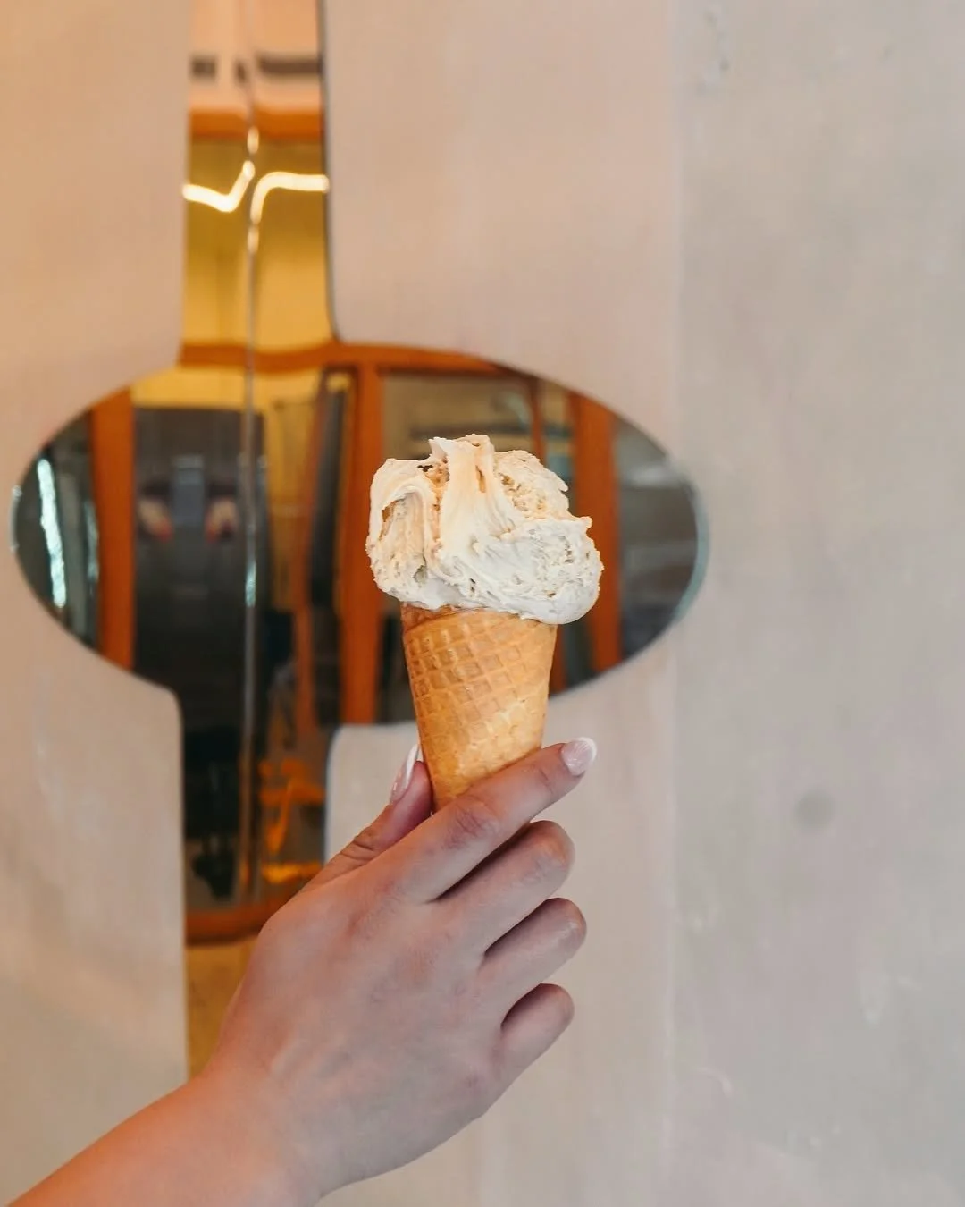 A hand holding a cone with vanilla ice cream against a mirror background.