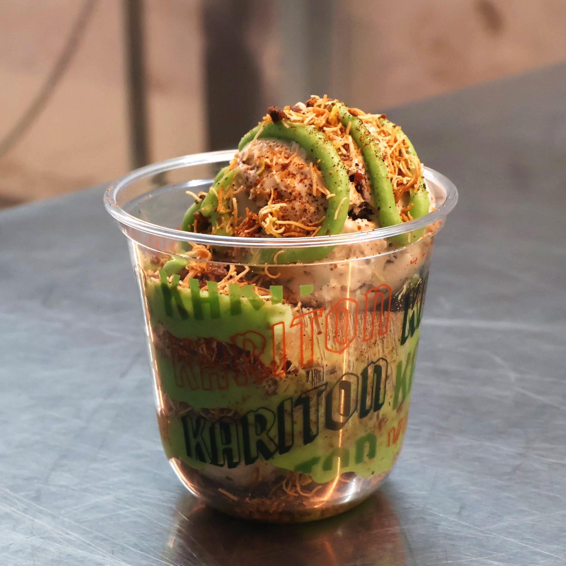 Ice cream dessert topped with crushed cookies, green gummy worms, and shredded cheese in a clear plastic cup on a metal surface.