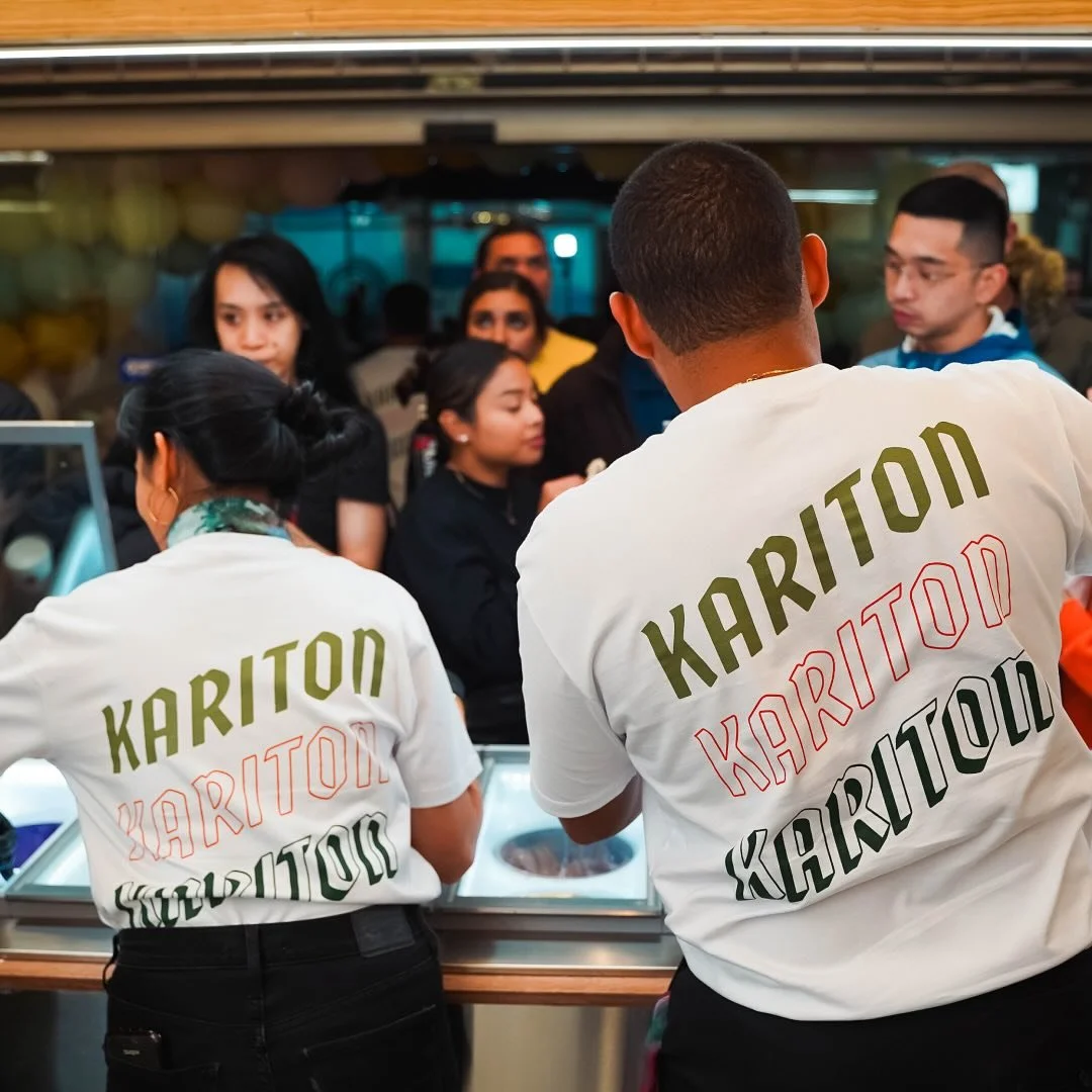 Two employees wearing white shirts with 'KARITON' printed on the back, working at an ice cream or food counter with customers in line behind them.
