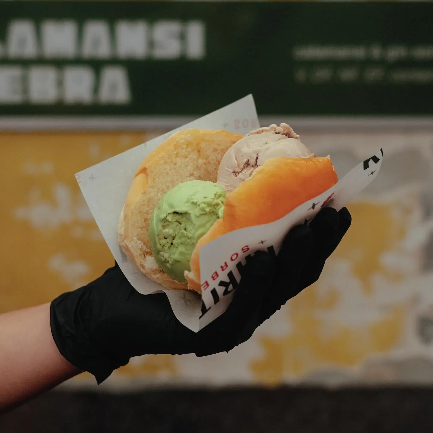 A hand wearing a black glove holding a tray of three scoops of gelato in waffle cones, with a blurred background.