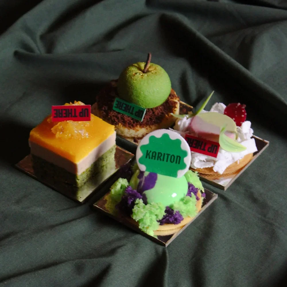 A tray of four decorated miniature desserts, including a green apple on a chocolate base, a layered treat with yellow and white layers and a red "UP THE RE" tag, a green and purple decorated dessert with a "KARITON" label, and a strawberry-topped pastry with whipped cream and a pink tag.