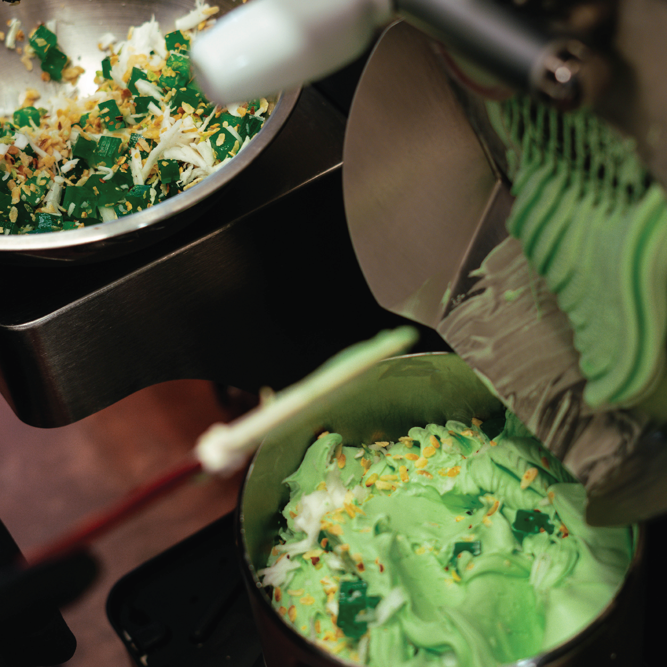 Ice cream being made with green ice cream in an ice cream machine, topped with sprinkles, next to a bowl of chopped green and white toppings.