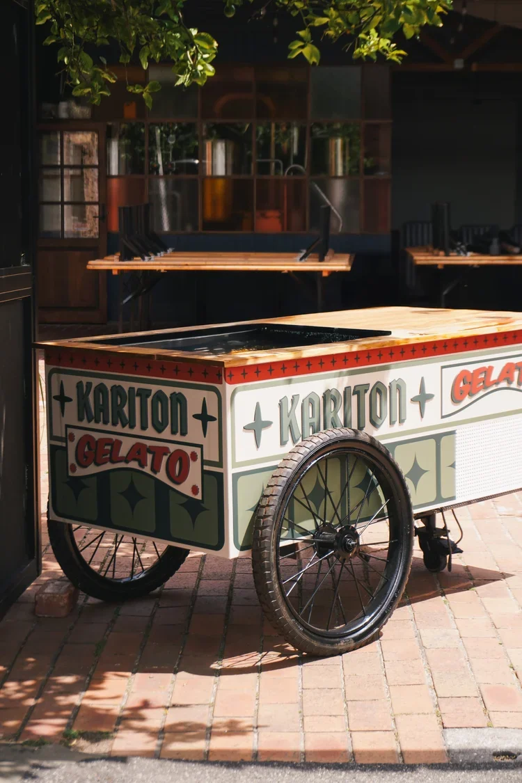 A vintage ice cream cart with the words 'KariTon Gelato' on the side, parked on a brick sidewalk in front of a building with green leaves hanging above.