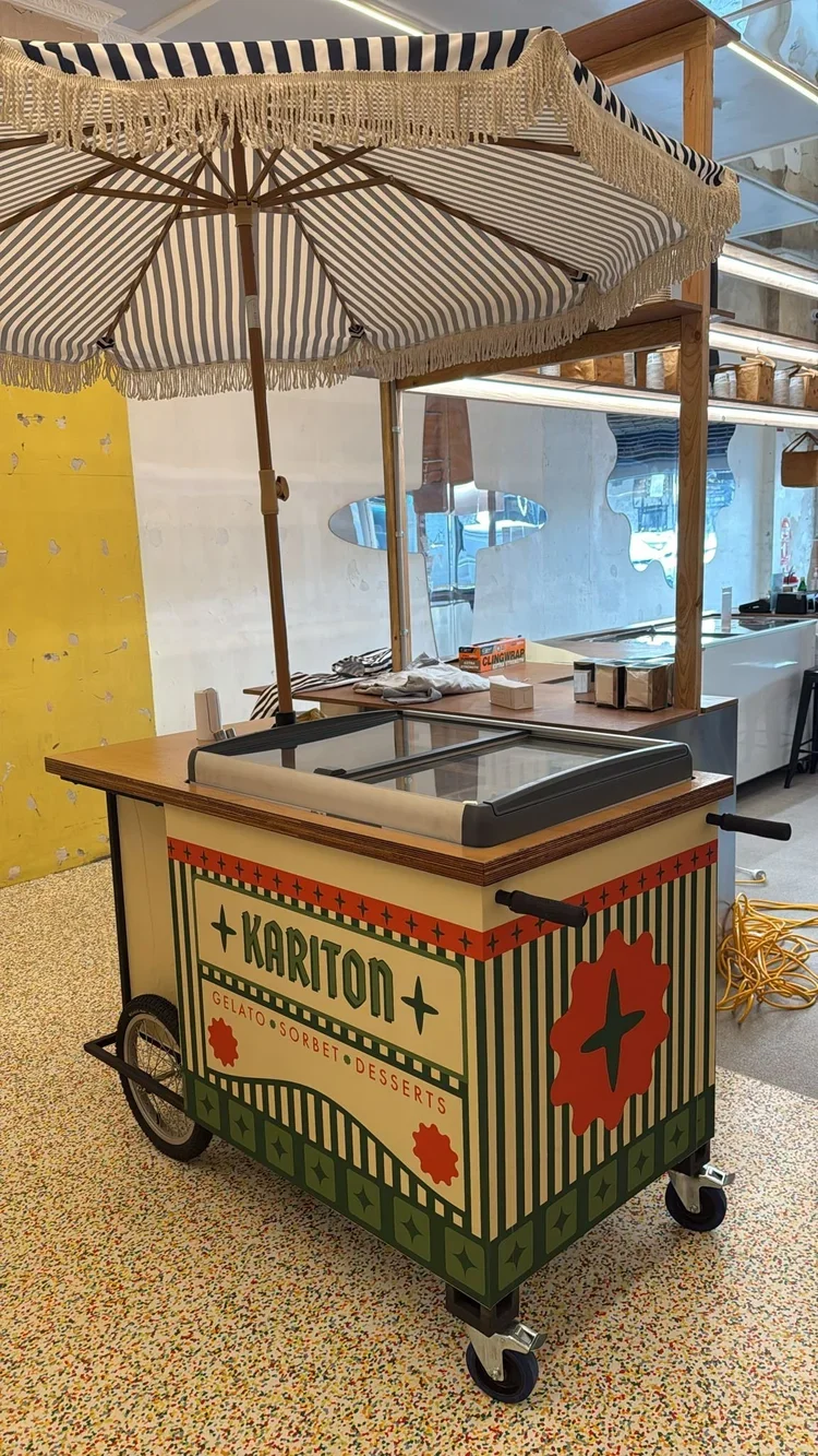 A vintage-style mobile ice cream cart with a striped umbrella, decorated in green, red, and cream colors, with the name 'KARITON' on the front, and options for gelato, sorbet, and desserts.