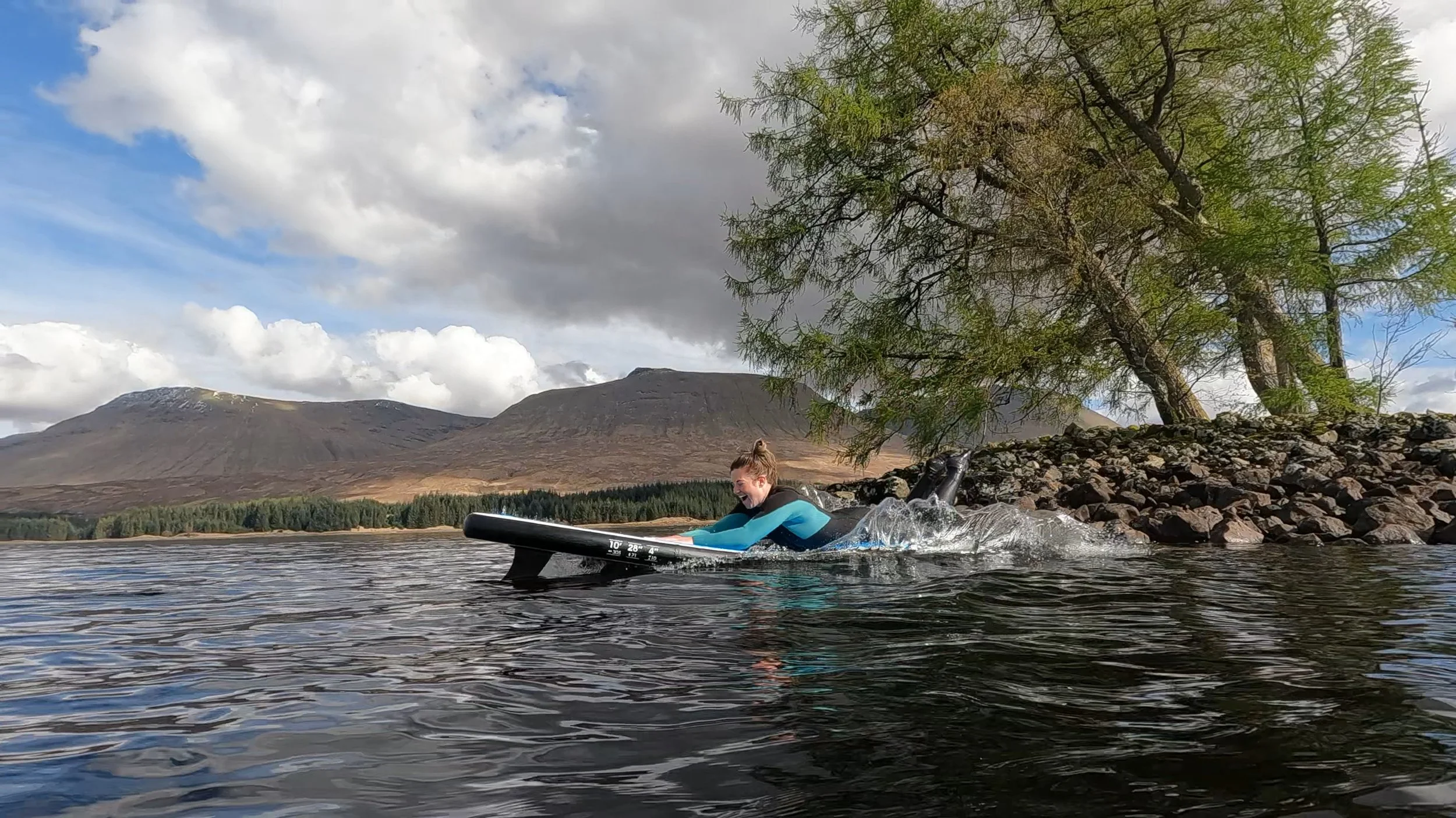 A person in a wetsuit riding a paddleboard on a loch with Bridge of Orchy mountains, trees, and a blue sky with clouds in the background in the Scottish Highlands.