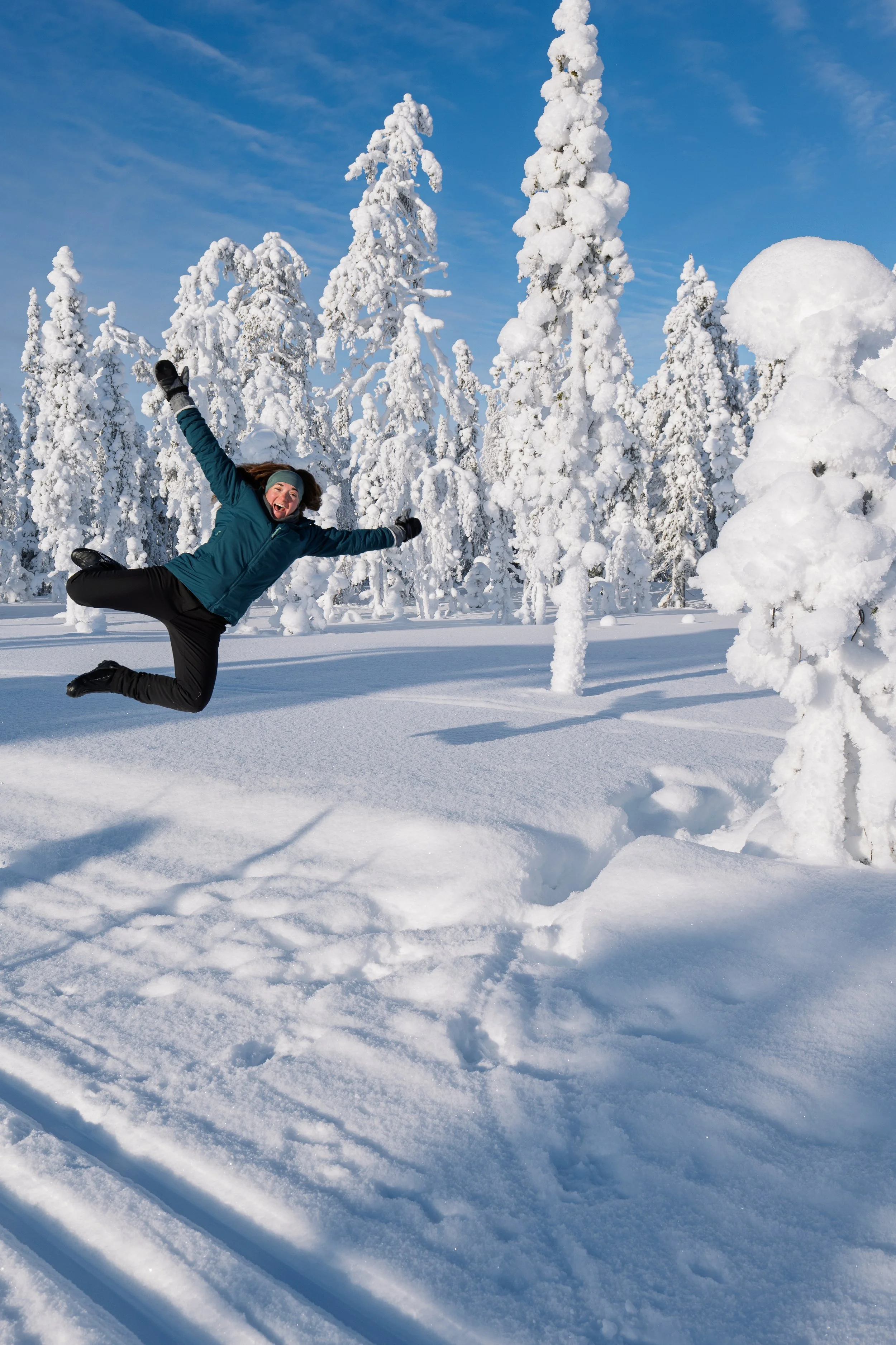A woman in winter clothing jumping joyfully in a snowy landscape with snow-covered trees and a clear blue sky in Finland about to land in the snow.