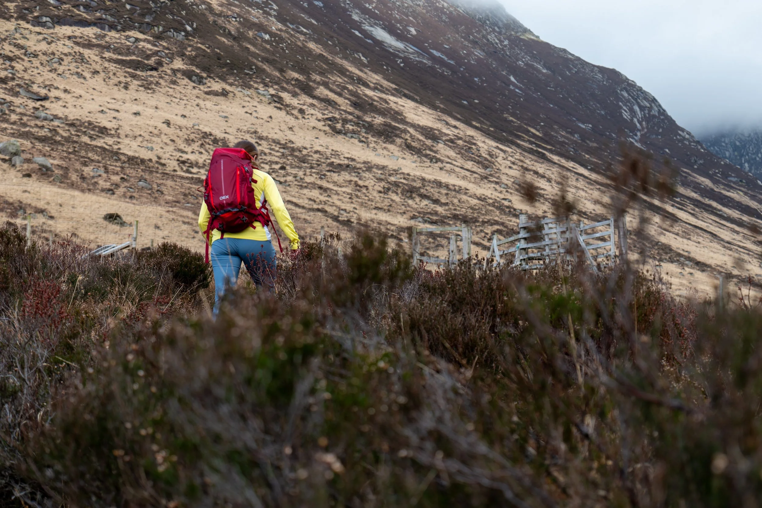 A female hiking in a mountainous area with a large red OEX backpack, yellow jacket, and blue pants, walking on a trail surrounded by dry vegetation and a mountain slope in the background of Isle of Arran.