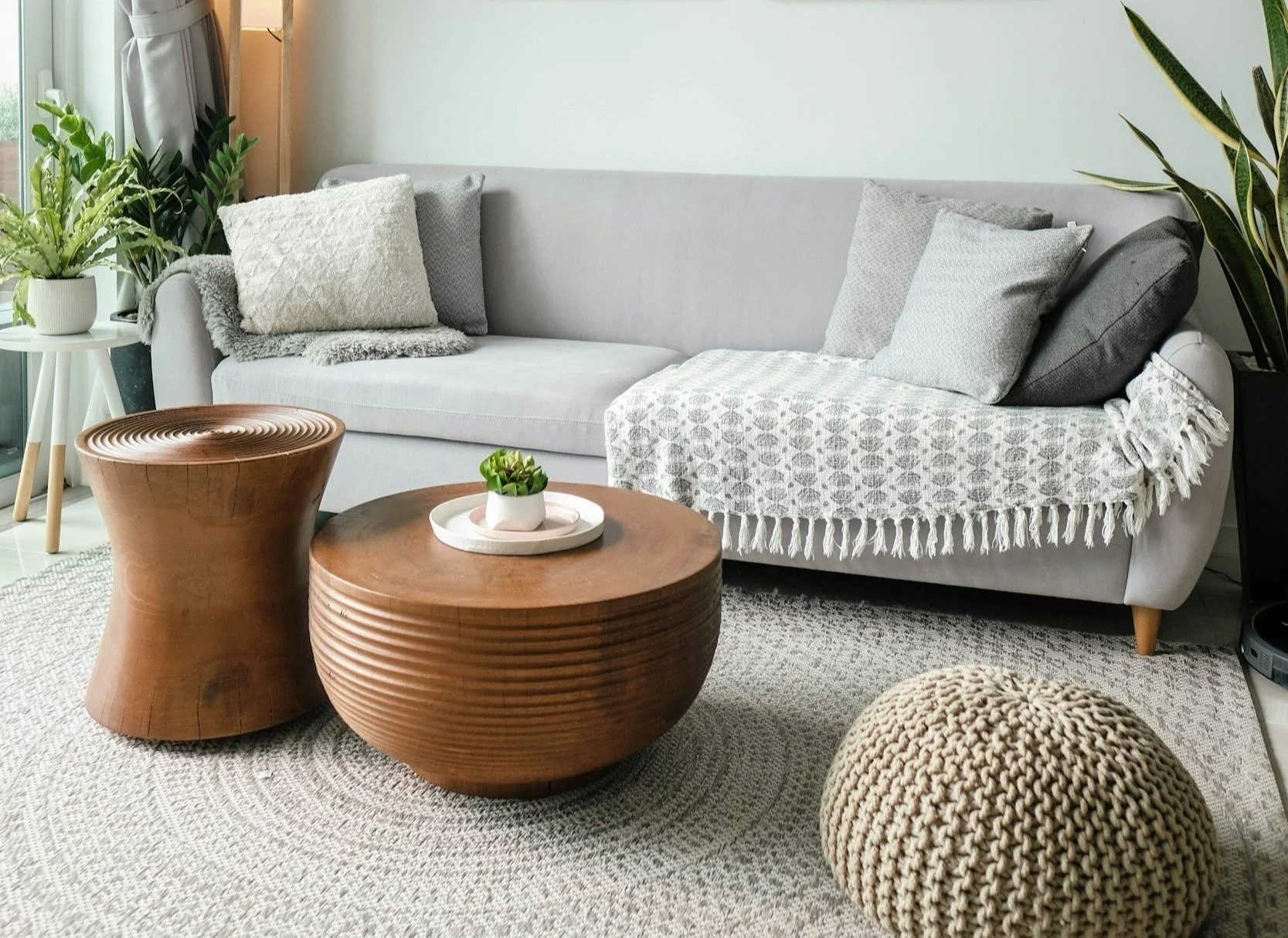 Living room with a light gray sofa, gray and beige throw pillows, and a white textured throw blanket. Two round wooden coffee tables, one with a small potted plant, are on a beige textured rug. Green plants in white pots are on a white side table and in a black pot at the end of the sofa. Natural light from a window illuminates the space.