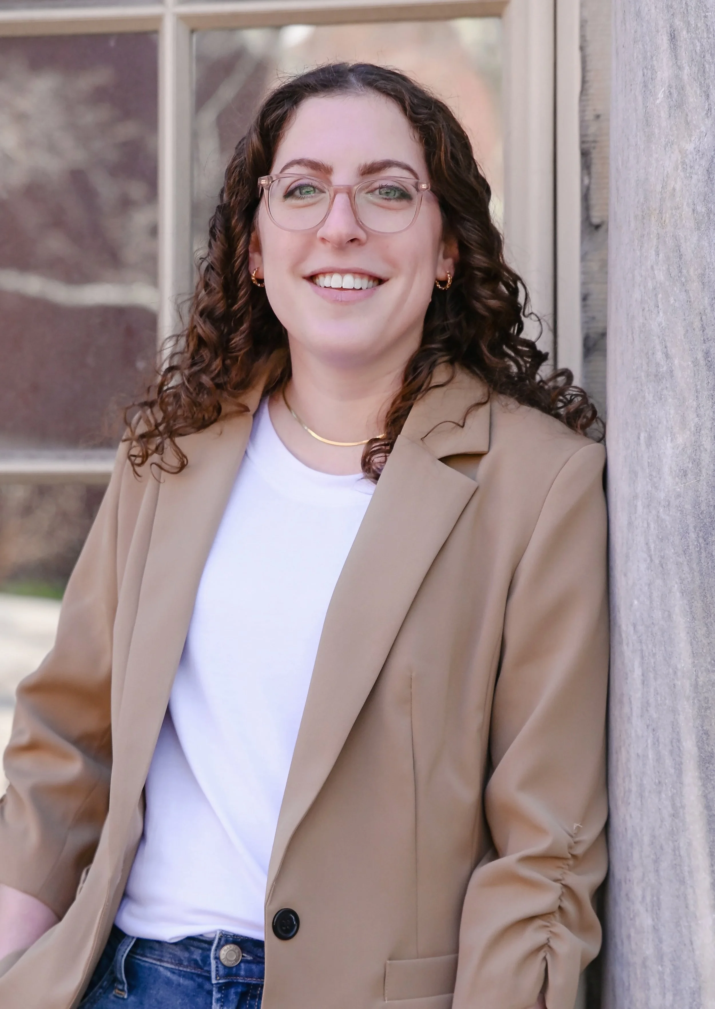 A young woman with curly hair, glasses, and gold jewelry smiling outdoors, leaning against a wooden wall.