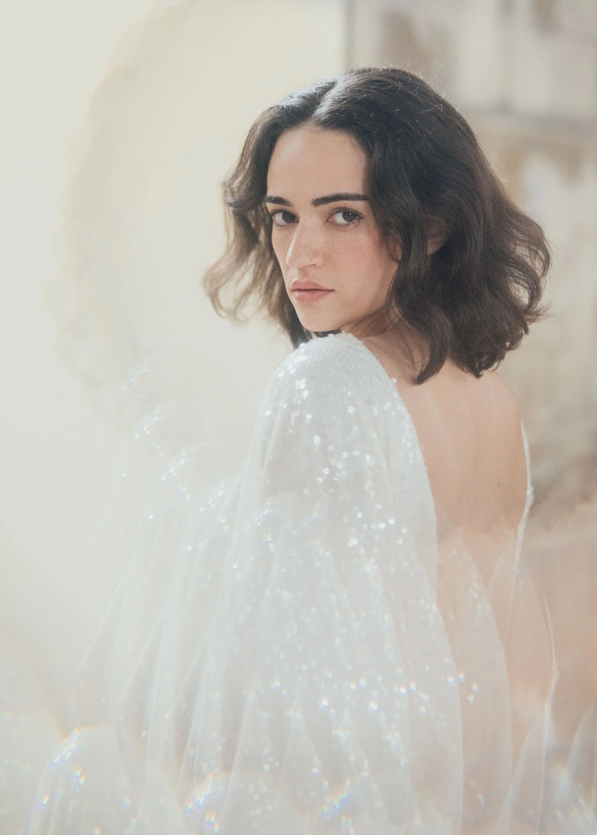 Ophelia Dress by Rolling in Roses – shimmering georgette bridal gown fully embroidered with subtle sequins, featuring a high neckline, plunging open back, fitted skirt with petite godet, and modest train. Includes optional ribbon tie for added securi