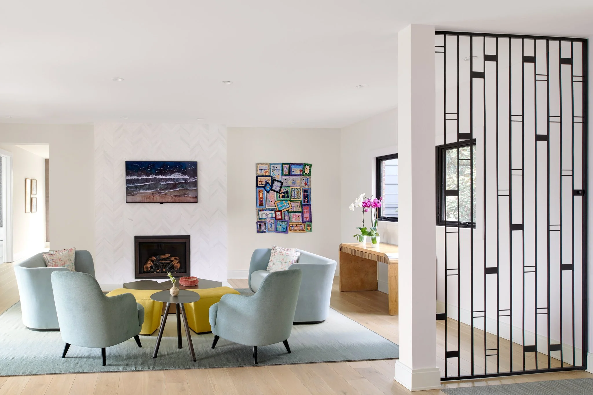 Luxury open-concept living room featuring a custom black metal geometric screen partition, honed white marble chevron tile fireplace, and curved powder blue settees.