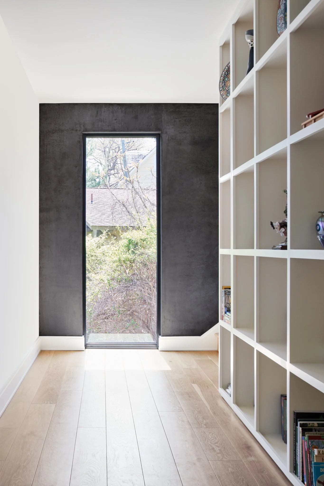 Minimalist hallway renovation featuring a floor-to-ceiling white gridded display bookcase, wide-plank oak flooring, and a dramatic dark textured plaster accent wall around a picture window.