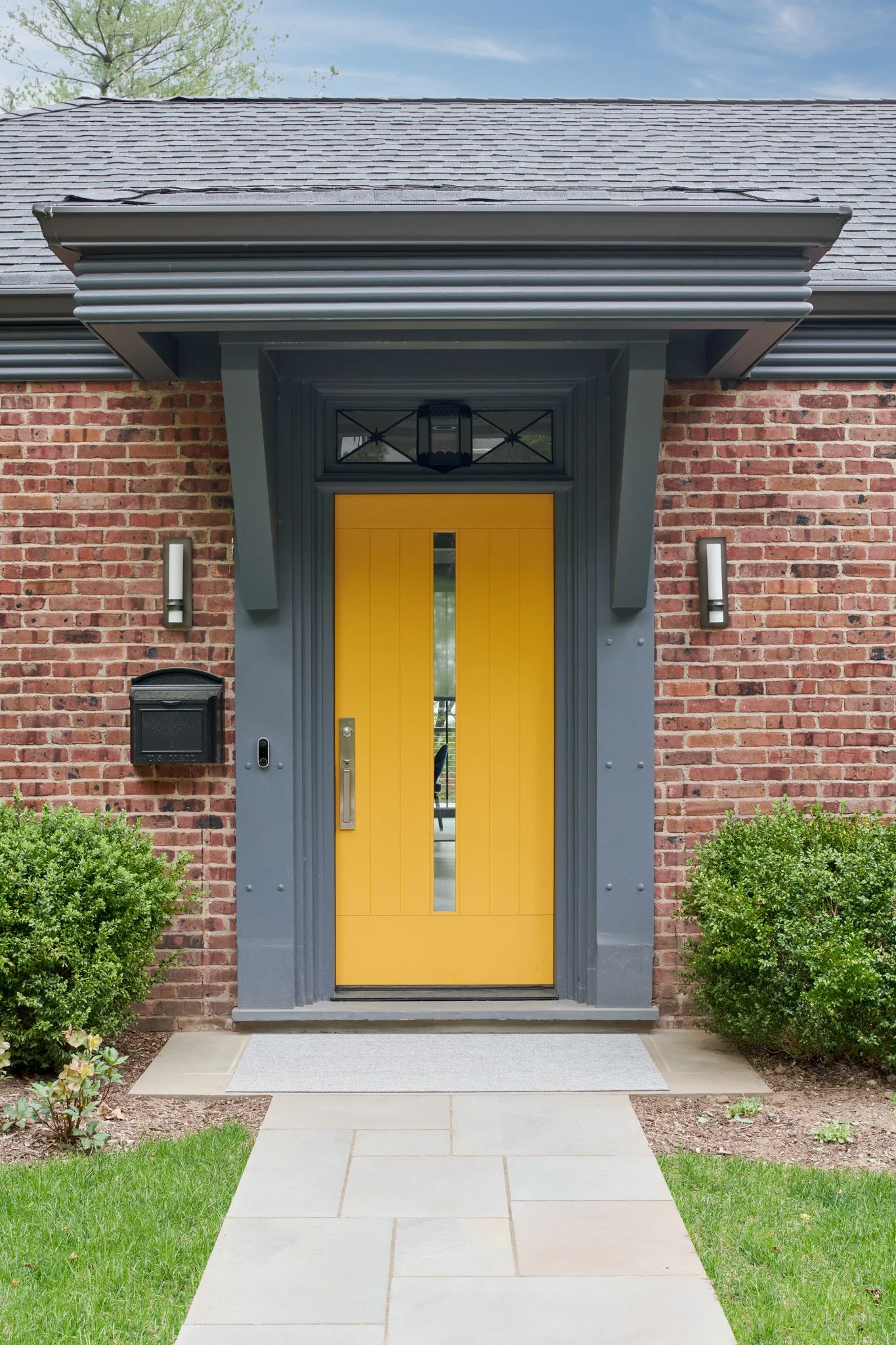 Mid-century brick ranch home transformed with a custom charcoal gray portico, structural brackets, modern sconces, and a vibrant yellow statement front door.