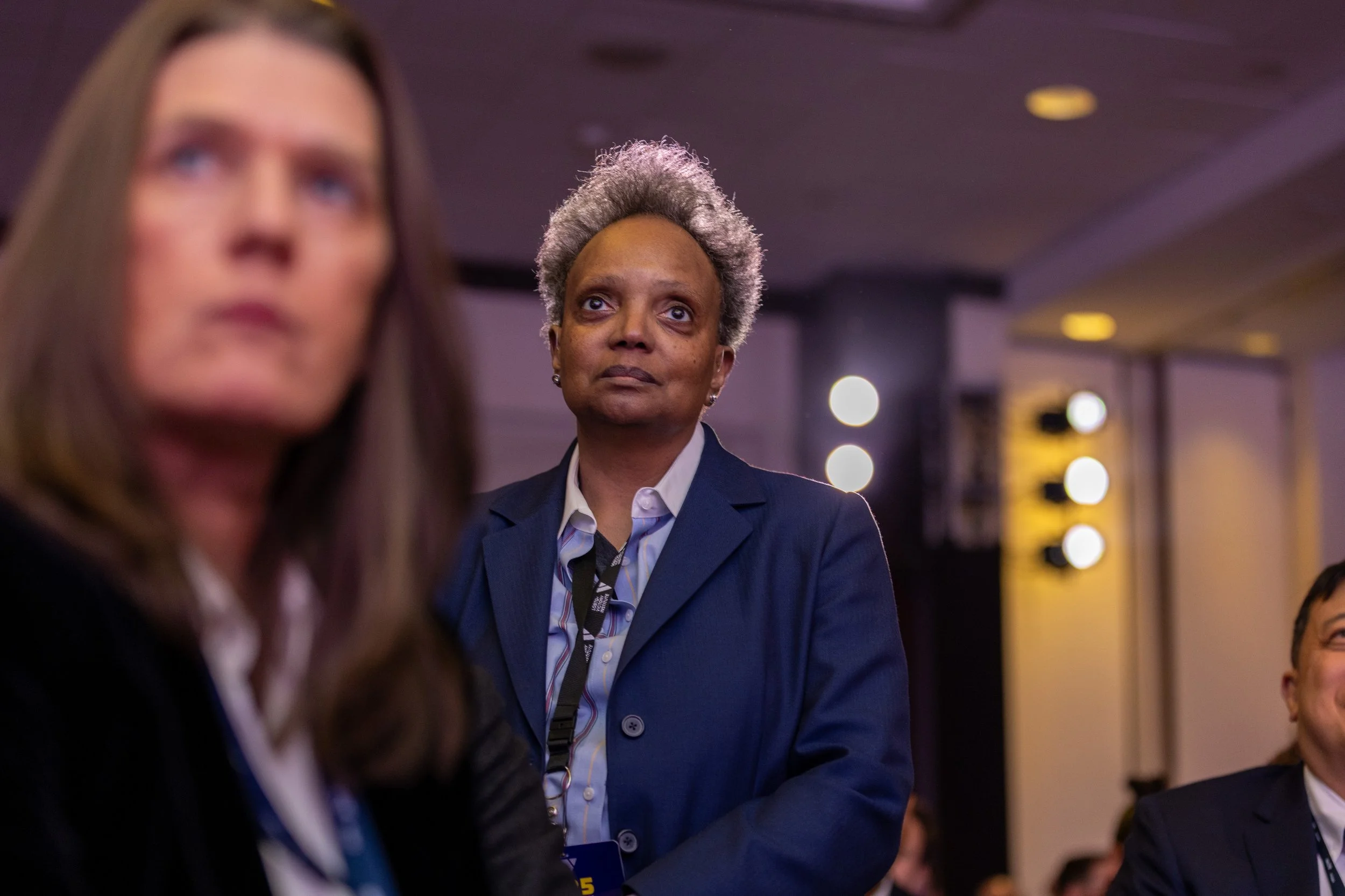 A middle-aged woman with short gray hair, wearing a navy blazer and white shirt, attentively listening at a conference or meeting in a large hall with stage lighting in the background.