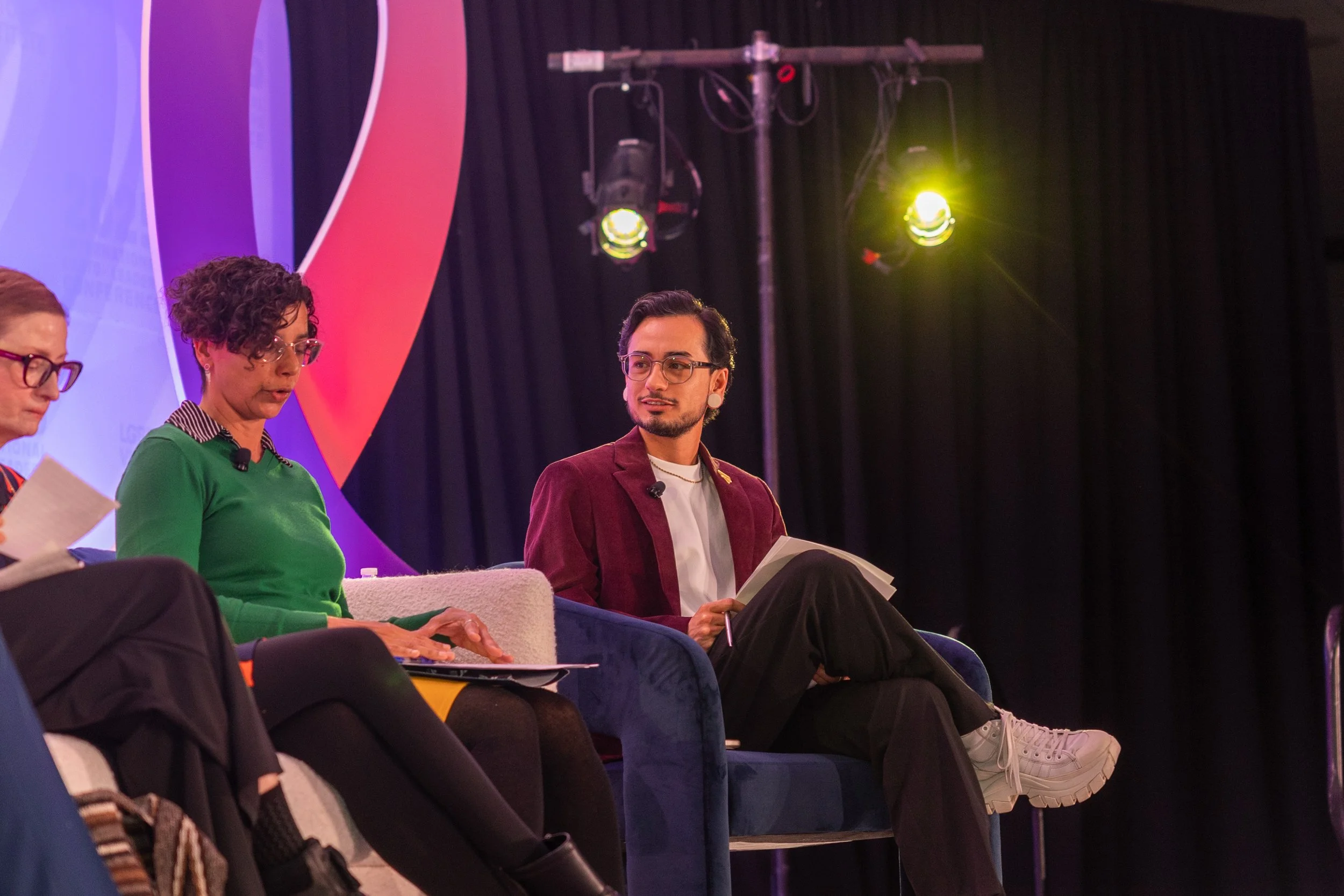 Panel discussion with three people on stage, including a man in a maroon blazer and glasses, a woman in a green sweater and glasses, and another woman on the far left. They are seated holding notes, with stage lighting and a large colorful backdrop b