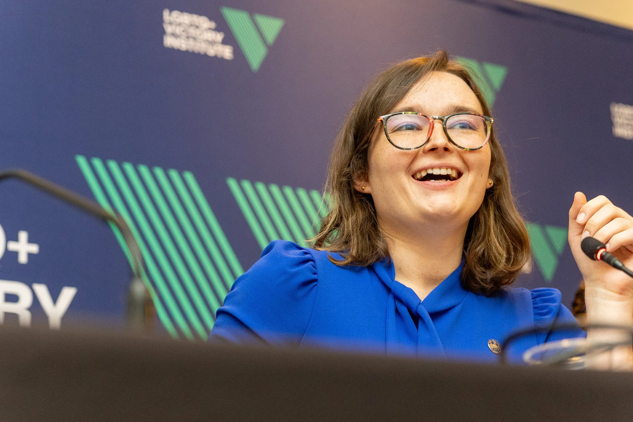 Young woman with glasses smiling at a conference, sitting in front of a backdrop with green and blue geometric designs and text.