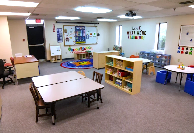 A bright, organized preschool classroom with child-sized tables and chairs, educational wall decorations, a colorful circular rug, toy shelves, and learning materials.