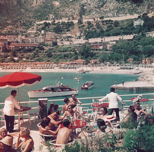 People dining outdoors at a seaside restaurant with a view of boats on turquoise water and a beach with buildings and greenery in the background.