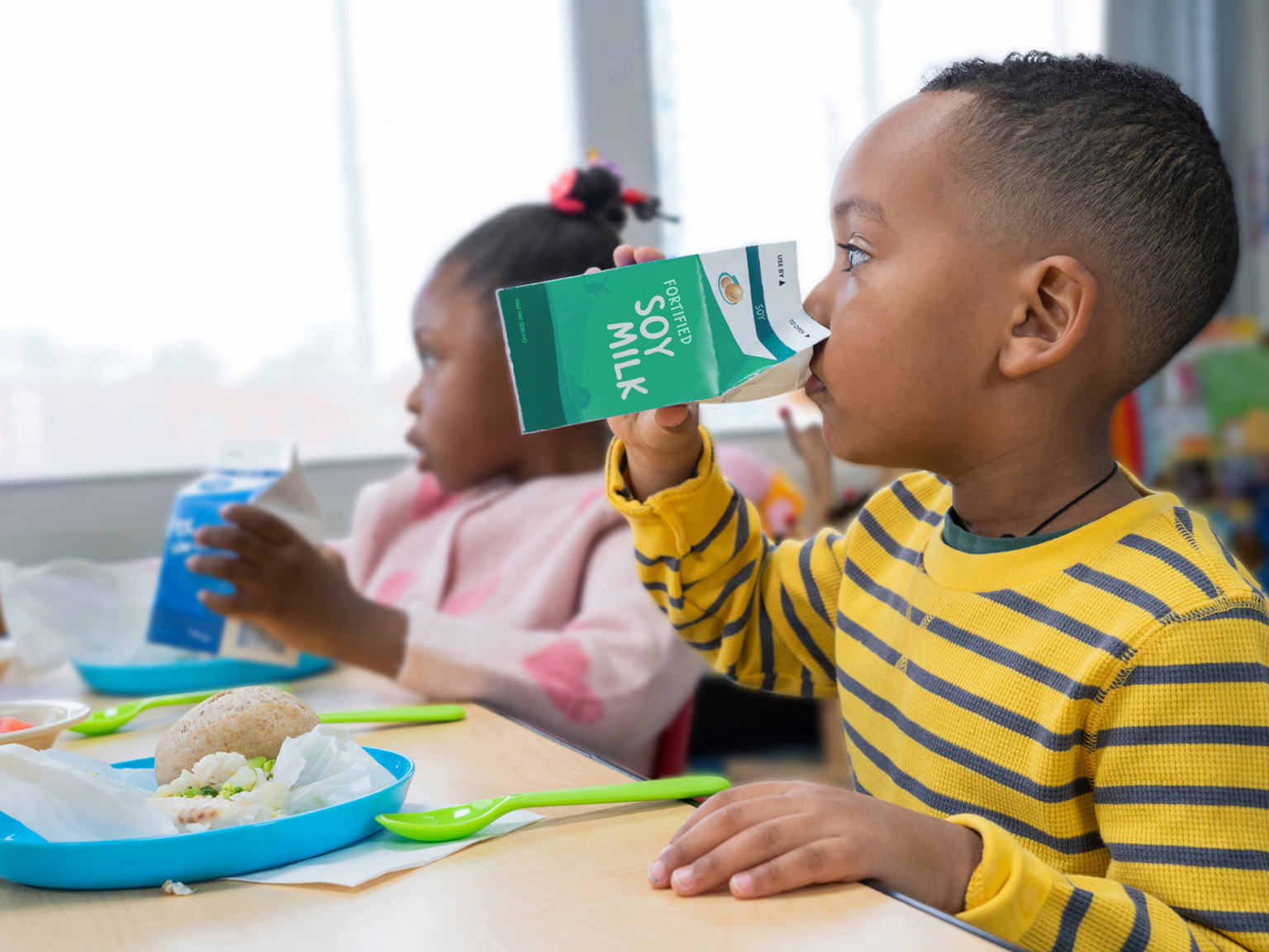 A young boy in a yellow and black striped shirt drinking soy milk from a green carton at a lunch table, with a girl in a pink sweater in the background also holding a carton.
