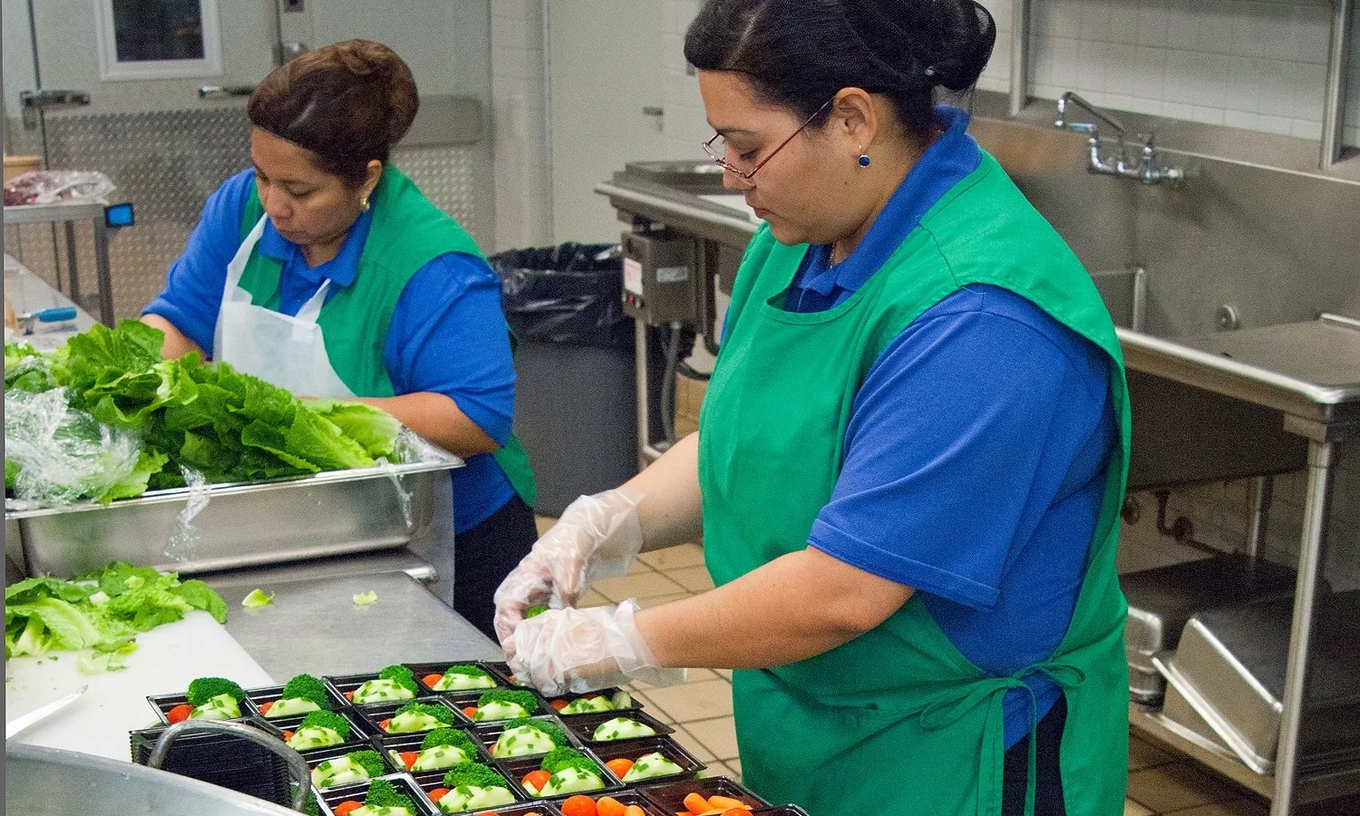 Two women in kitchen preparing food, with one arranging vegetable trays and the other slicing vegetables, both wearing blue shirts, green aprons, and gloves.