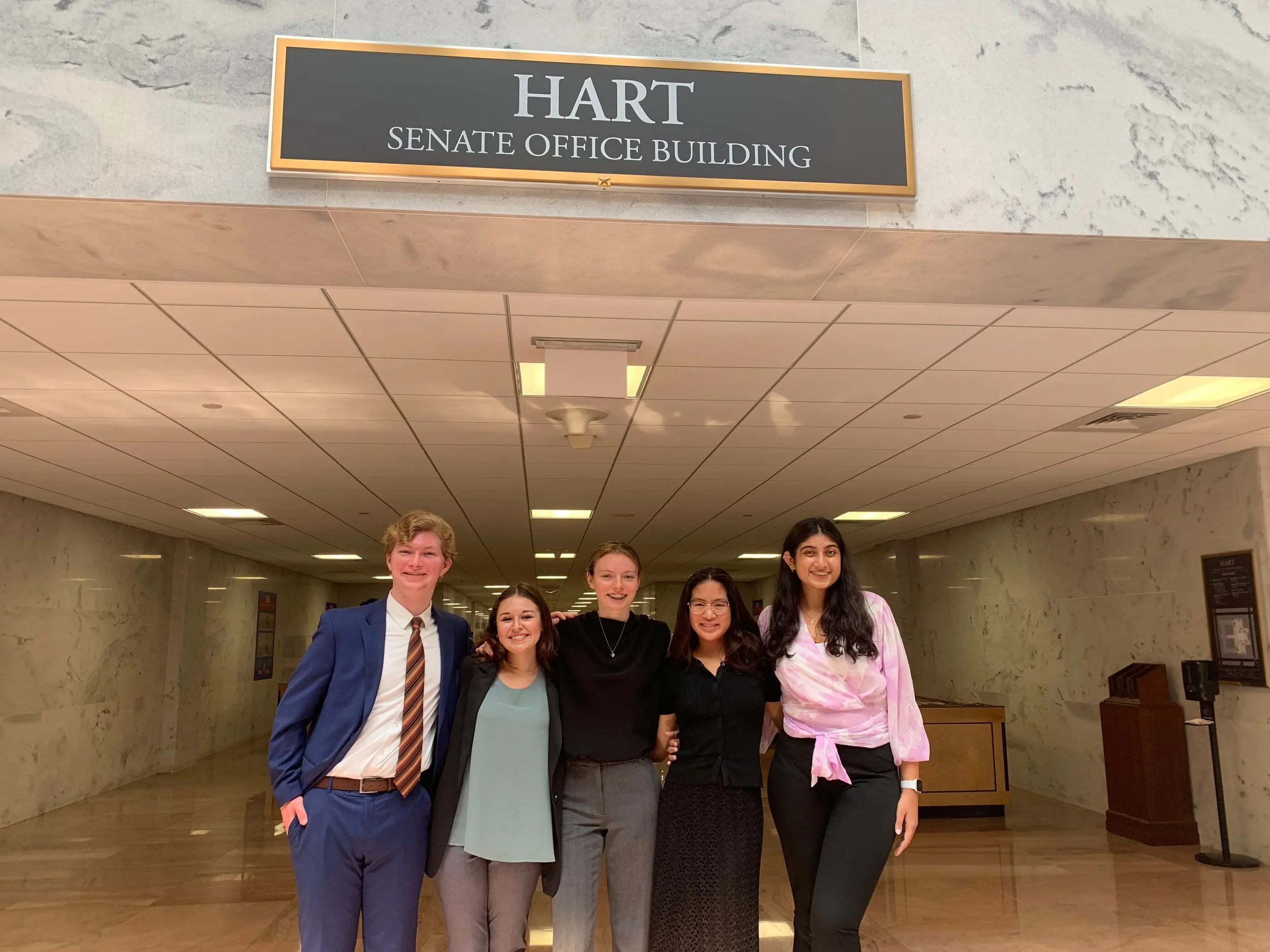 Group of five young adults, three women and two men, standing arm-in-arm inside a government building lobby, with a sign overhead reading 'HART SENATE OFFICE BUILDING'.
