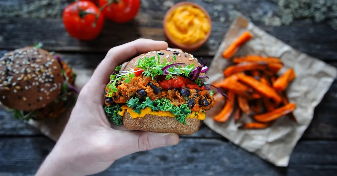 A hand holding a burger with various toppings, a side of sweet potato fries on parchment paper, a bowl of sweet potato mash, and fresh tomatoes on a rustic wooden surface.