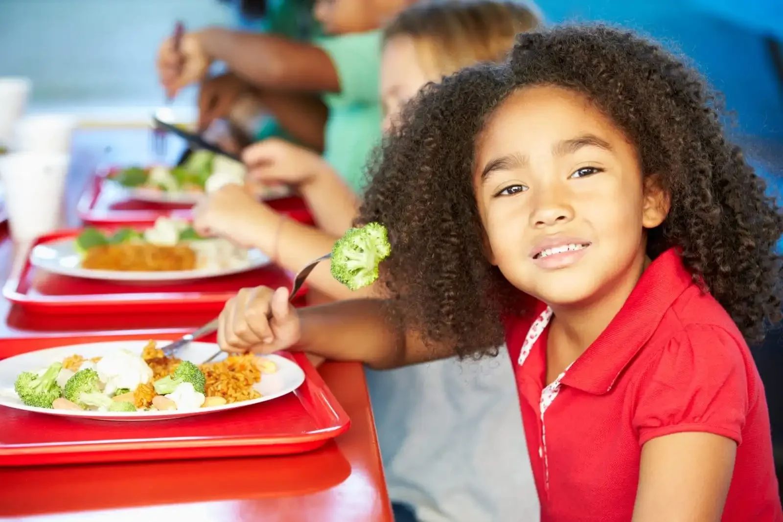 A young girl with curly hair and a red shirt eating food from a plate with broccoli, rice, and plant-based food while sitting at a long table with other children during a meal.
