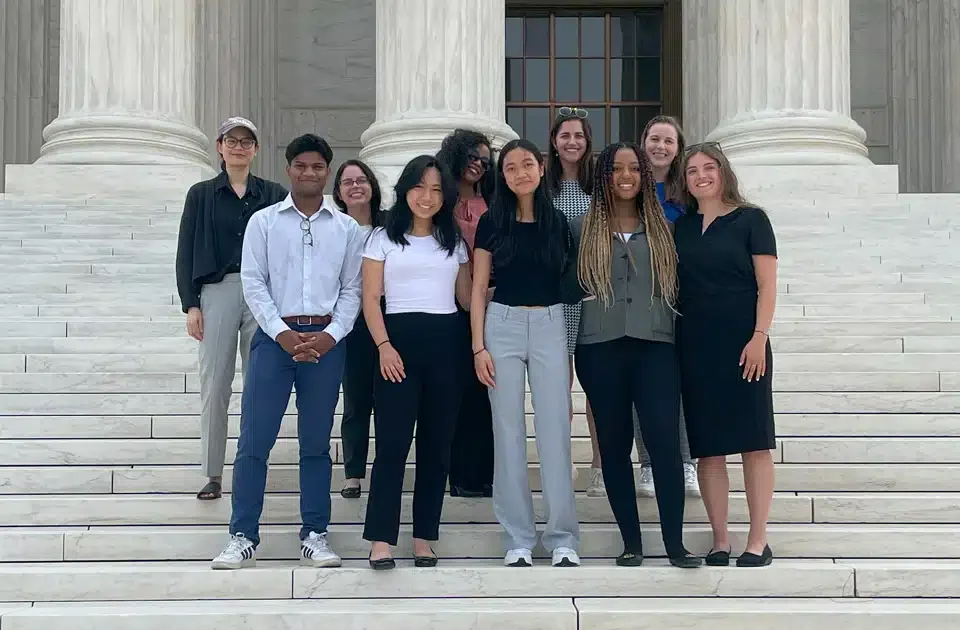Group of nine young women standing on the steps of a U.S. Supreme Court with large white columns, smiling at the camera.