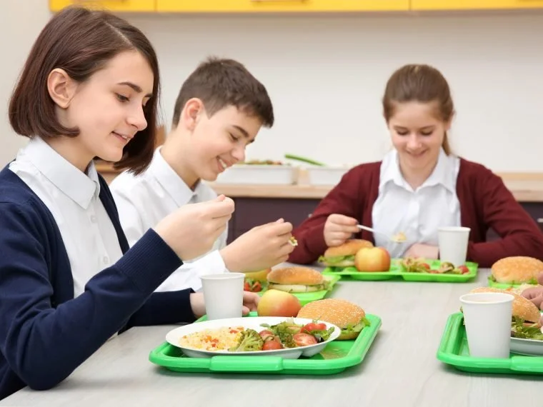 Three students having plant-based lunch at school, enjoying sandwiches, salads, and fruit, seated at a table with green trays.