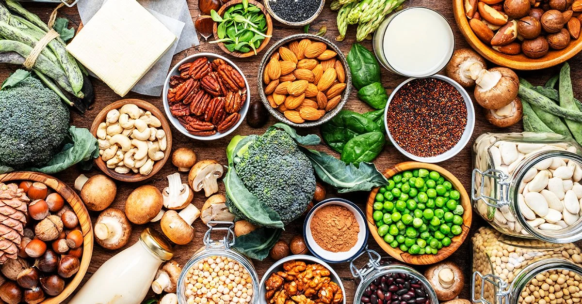 Assorted fresh vegetables, nuts, seeds, and grains displayed on a wooden table, including broccoli, mushrooms, almonds, cashews, walnuts, peas, lentils, and various spices.