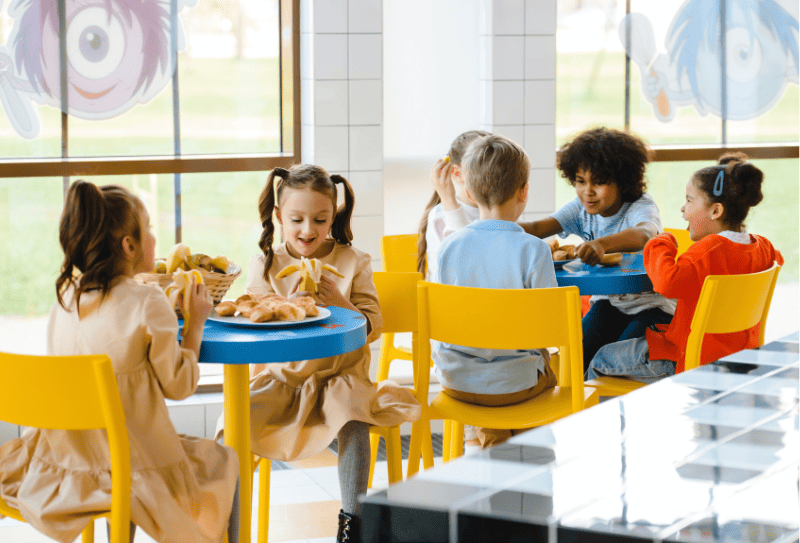 Children sitting at colorful tables in a brightly lit cafeteria, eating plant based snacks and chatting.